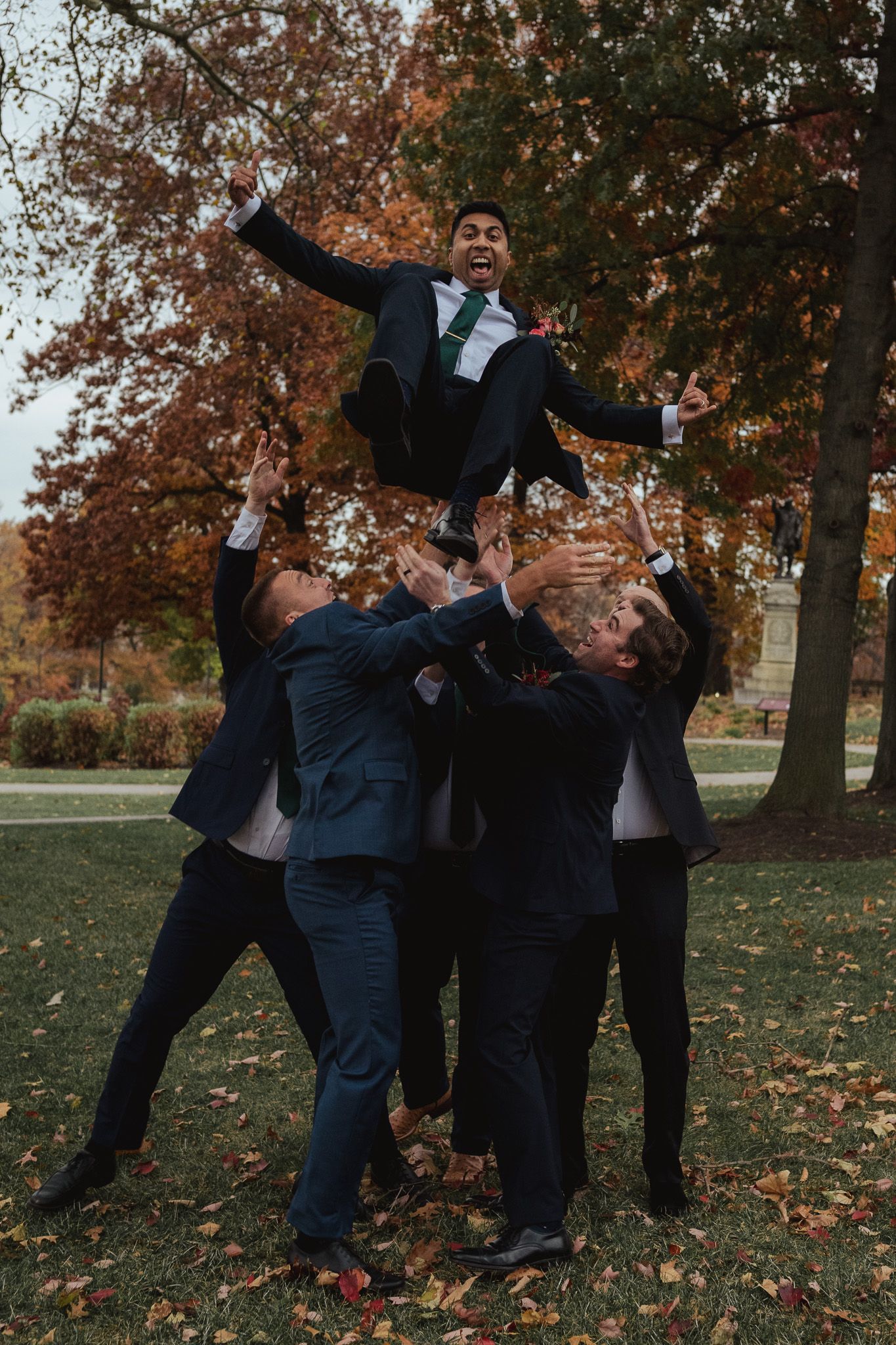 Group of people in formal attire playfully posing together in autumn setting with fallen leaves and trees.
