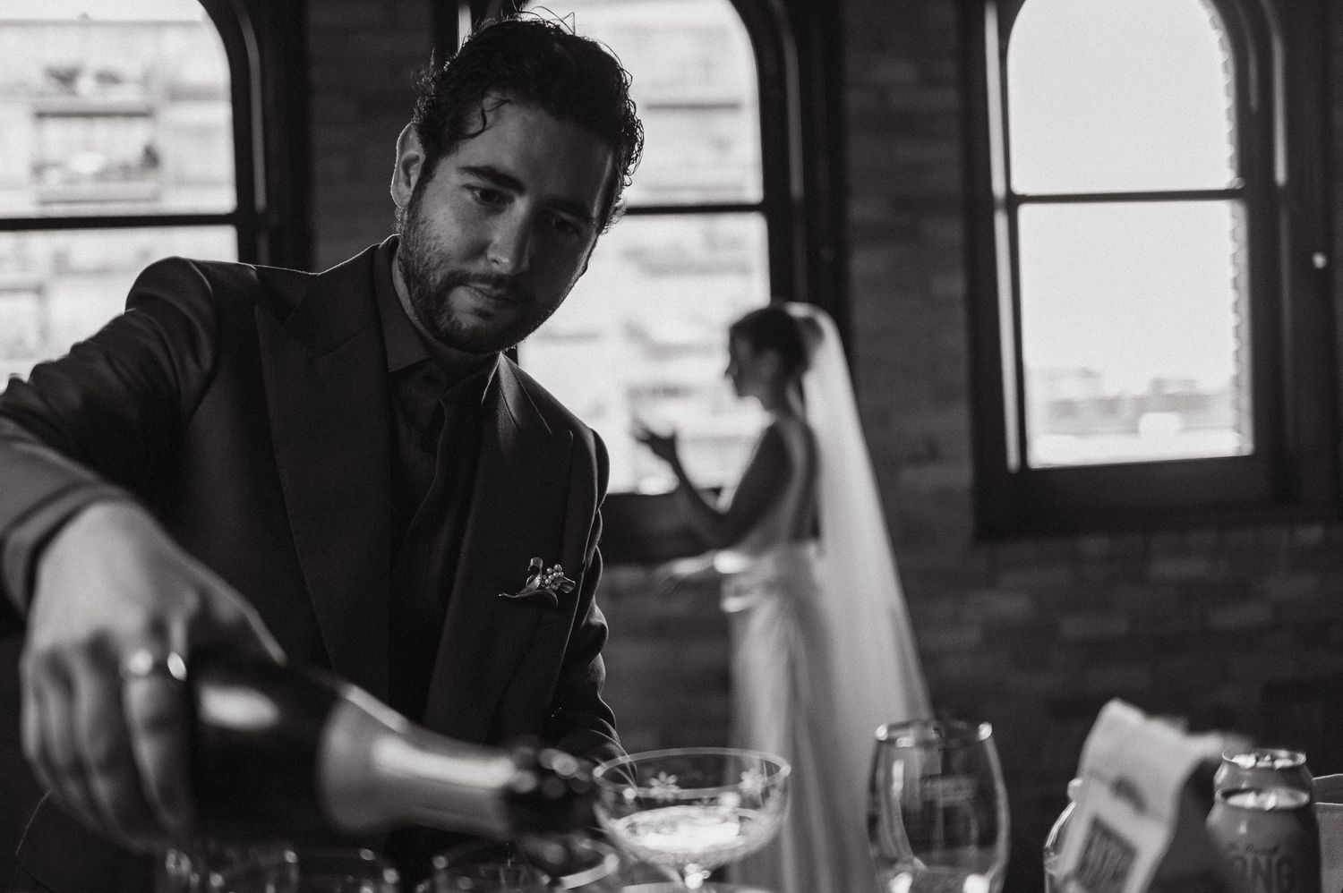 Black and white wedding photo of a man pouring champagne with a bride in the background at an elegant venue.