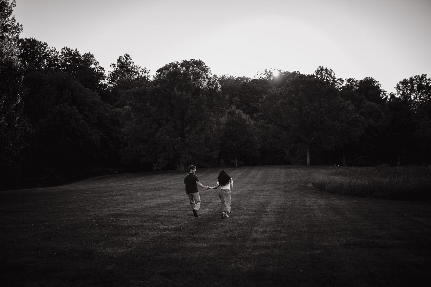 Two children hold hands while walking through a vast open field surrounded by lush trees at dusk in black and white.