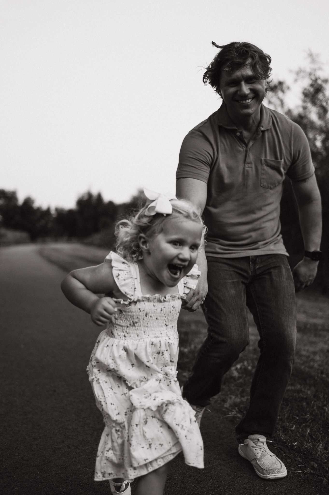 Black and white photo of a laughing little girl in a dress running with a smiling man on a road.