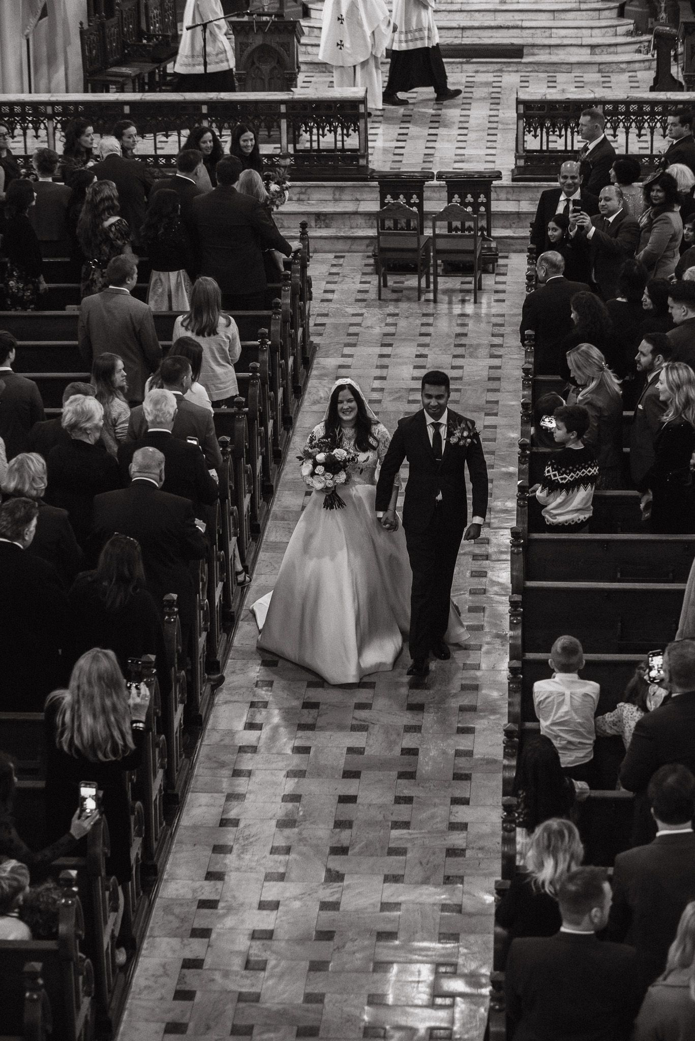 A black and white photo of a wedding ceremony in a grand church with guests seated in wooden pews.