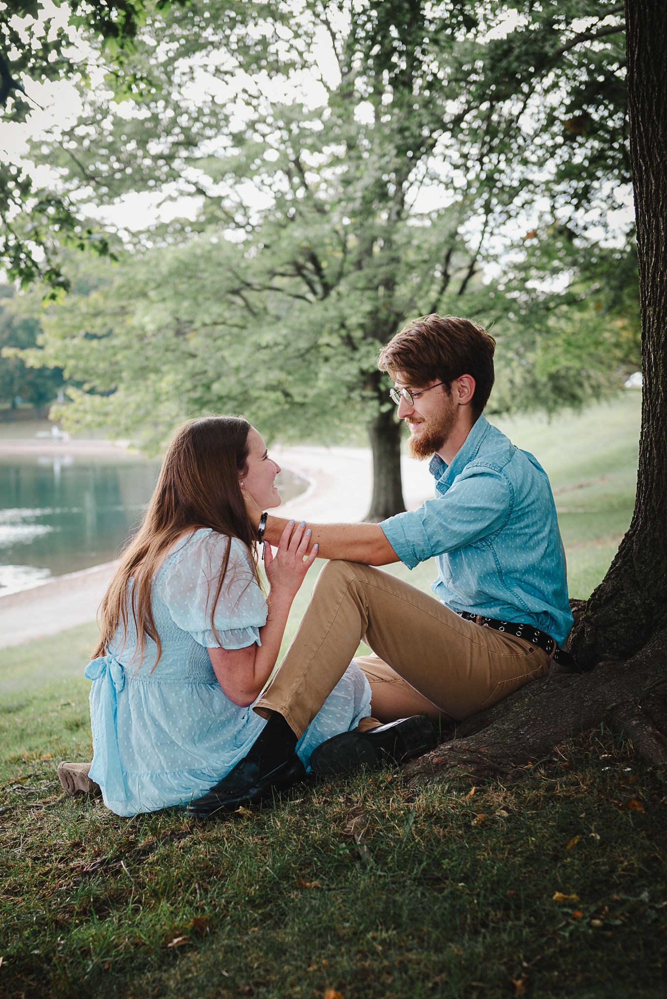 A romantic couple relaxes together under a shady tree by a lake on a summer afternoon.