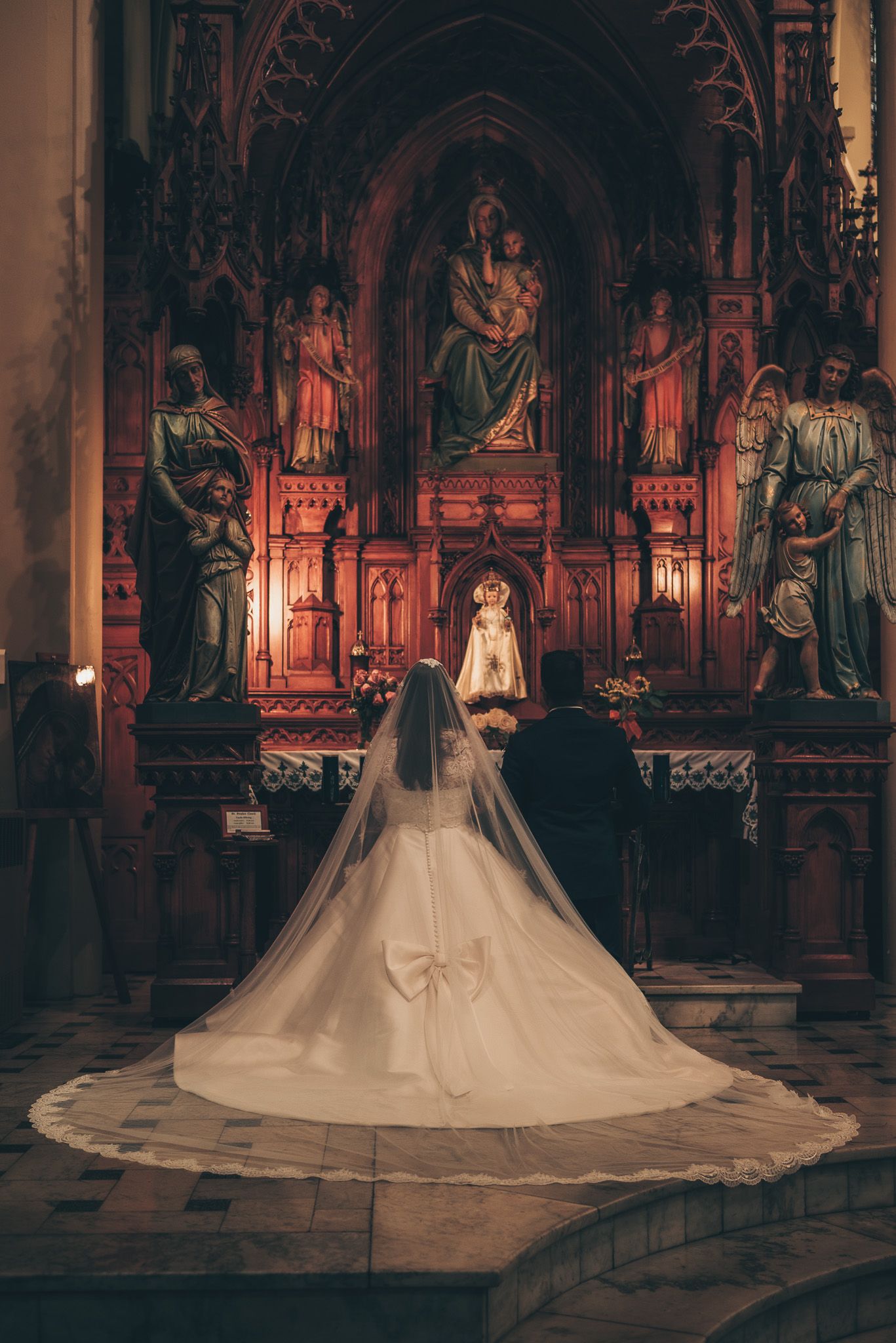A bride in a flowing white cathedral-length veil and gown stands before an ornate Gothic church altar with religious statues.