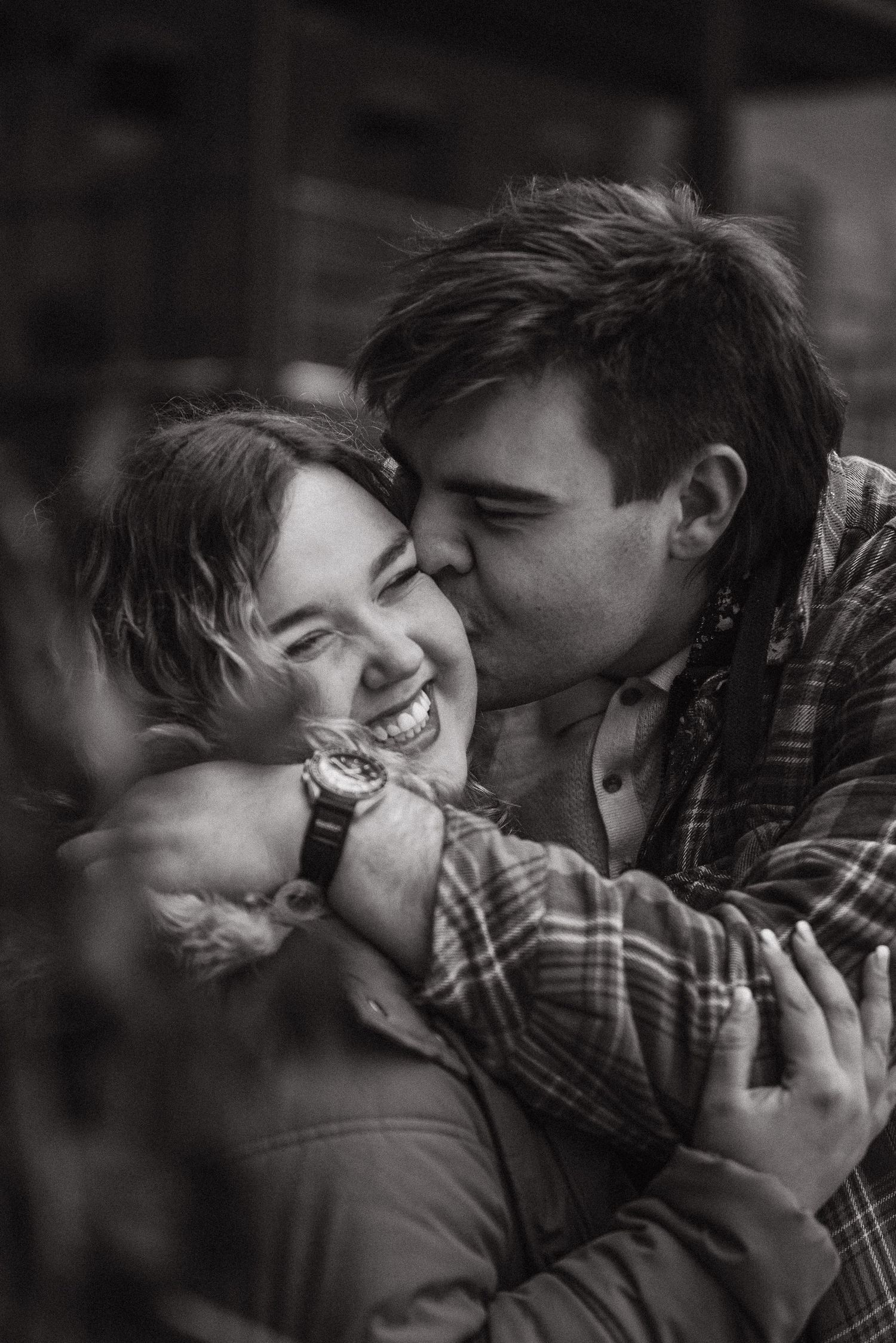 Black and white photo of a smiling woman being hugged and kissed on the cheek by a man wearing a plaid shirt.