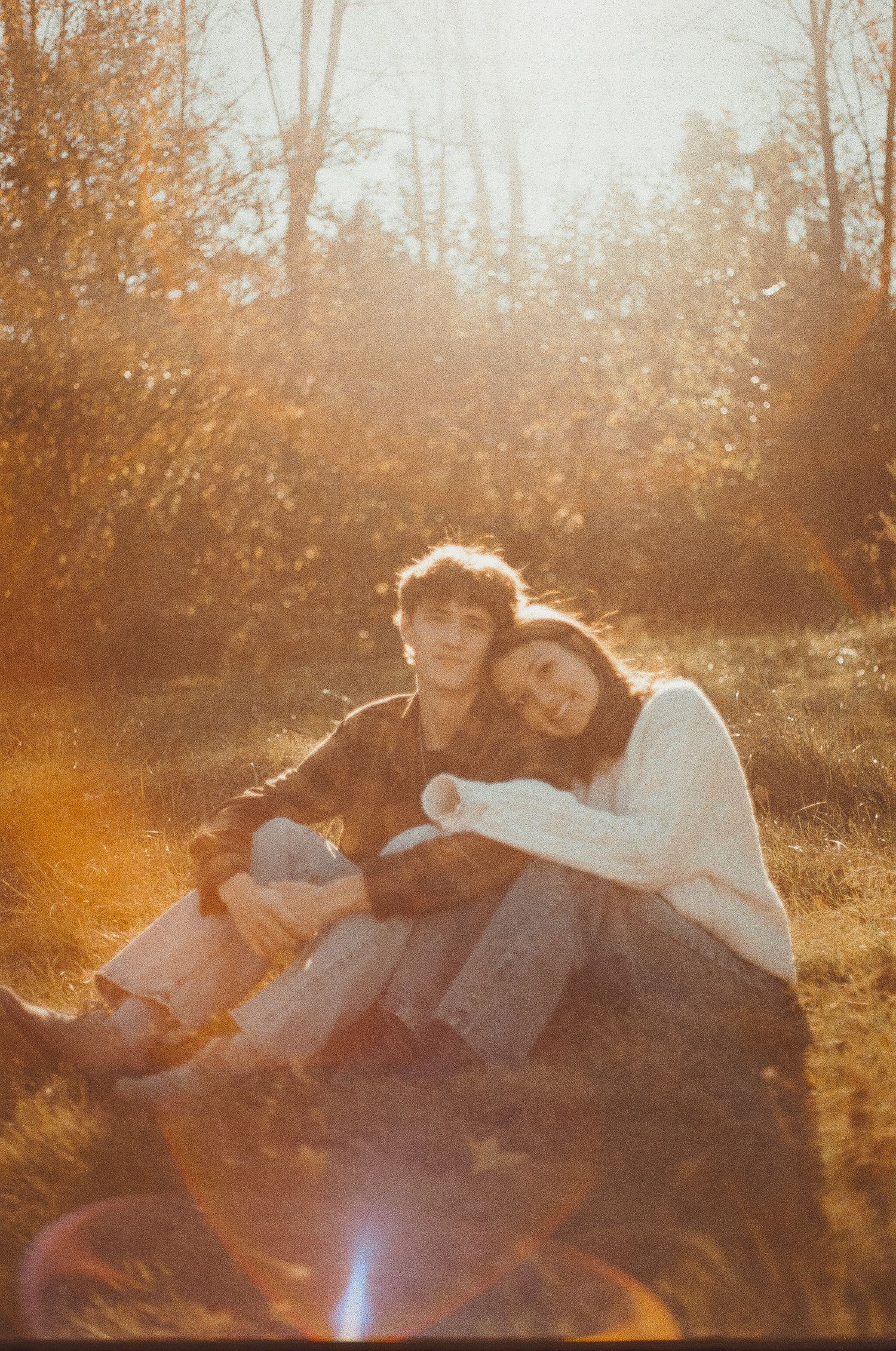 A couple lies together in a sunlit autumn field with golden light streaming through trees creating a dreamy atmosphere.