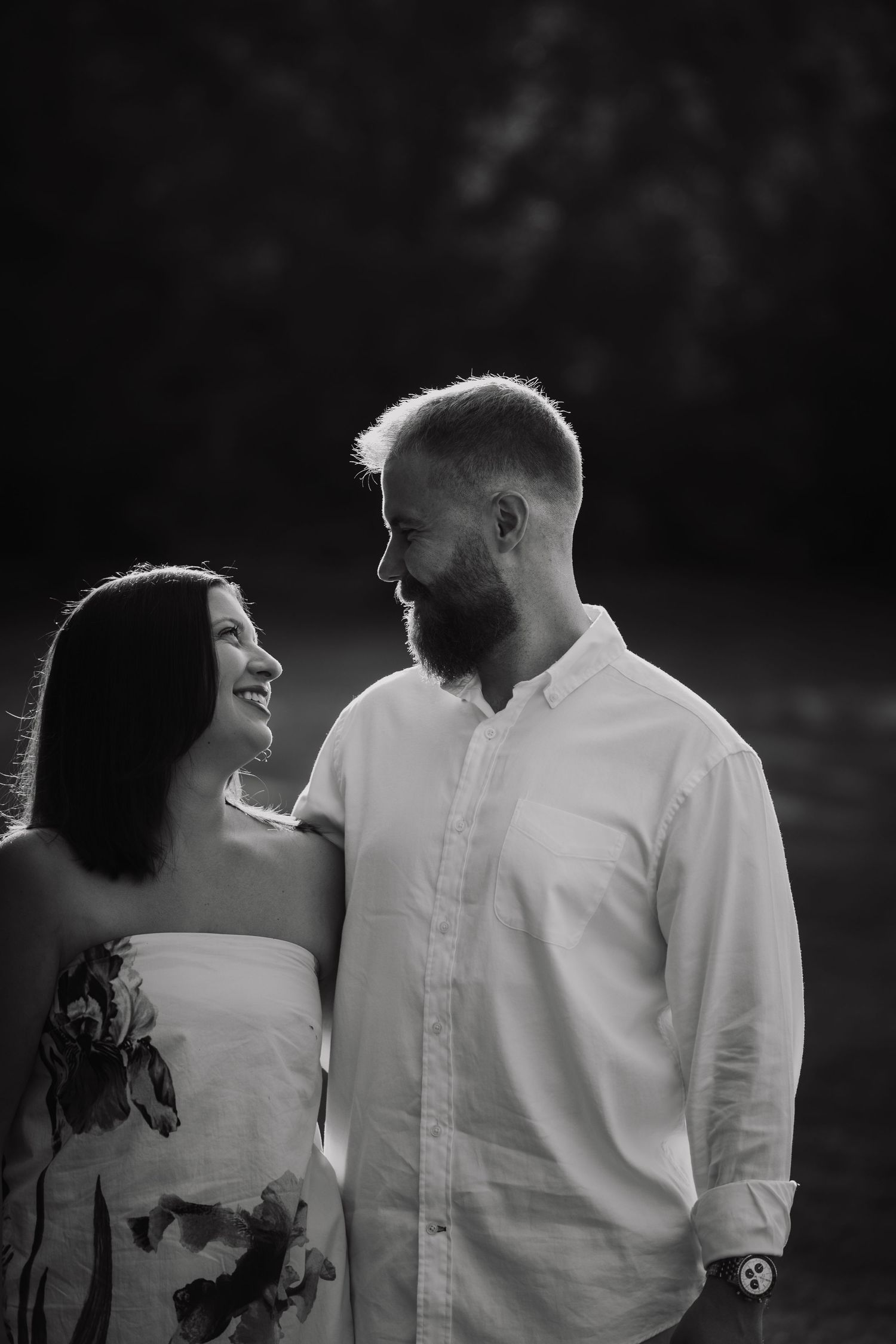 Black and white photo of a smiling couple gazing at each other outdoors, woman in floral dress, man in white shirt.
