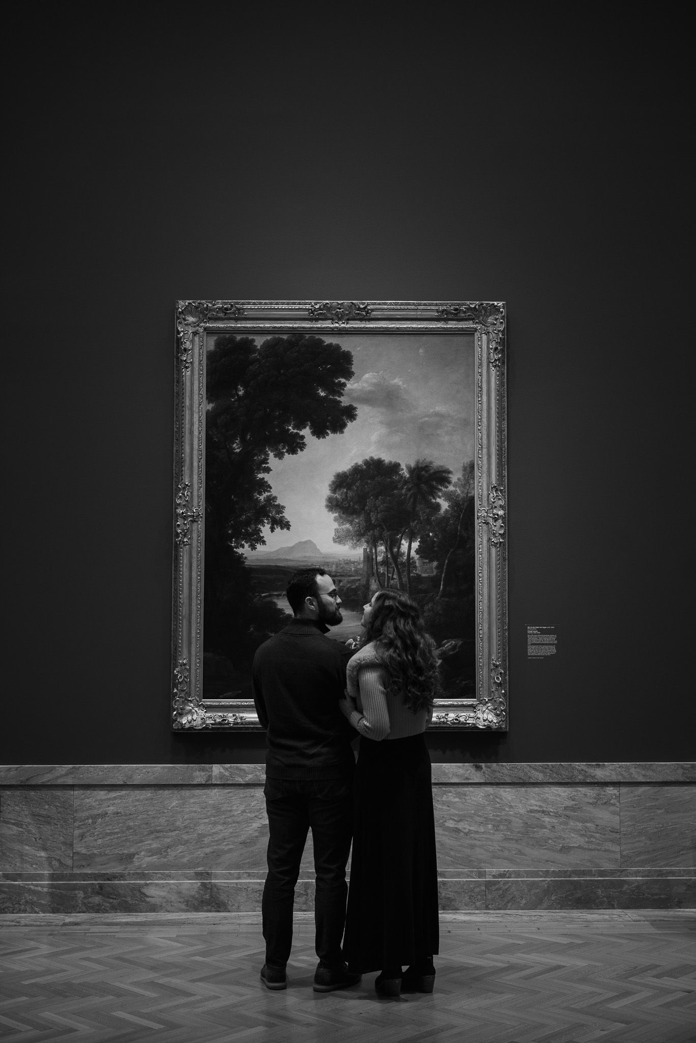 Silhouettes of visitors admiring framed landscape paintings in a dimly lit art gallery.