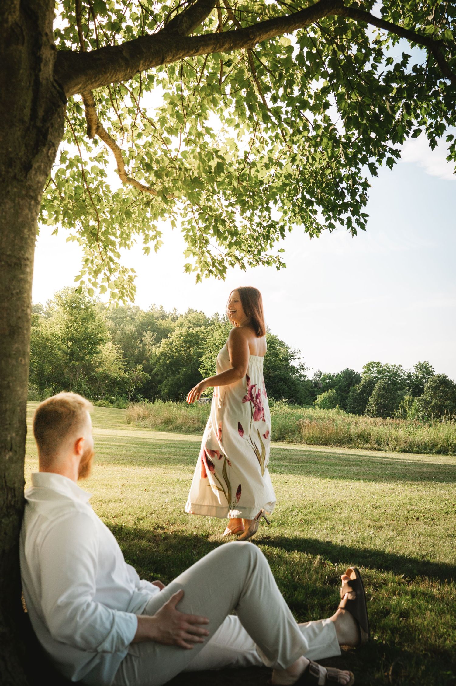 Couple in golden hour sunlight under a tree, woman in floral dress standing while man relaxes on grass in park.