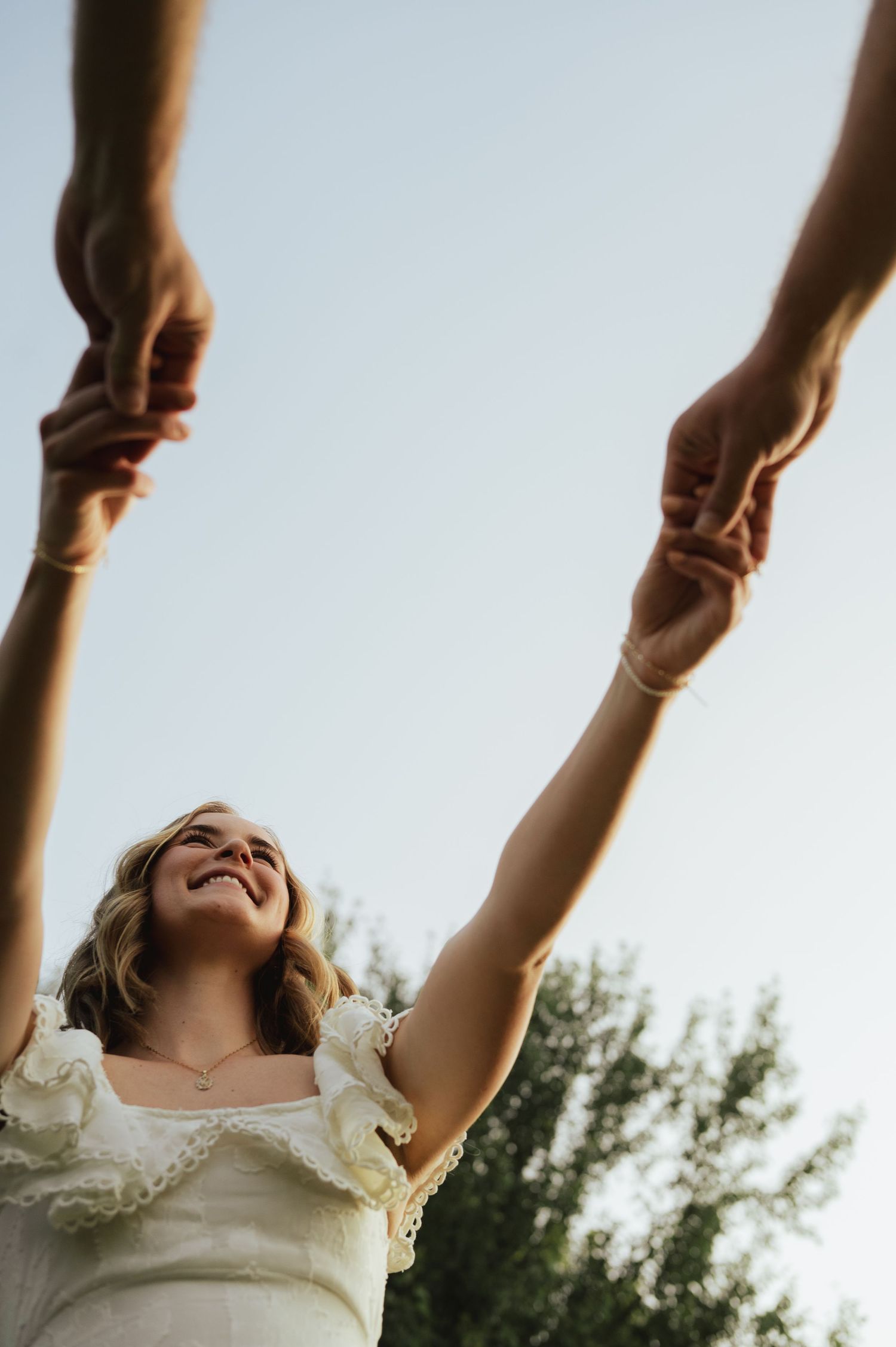 Smiling woman in white dress holds hands raised toward sky, photographed from below against a light blue sky.