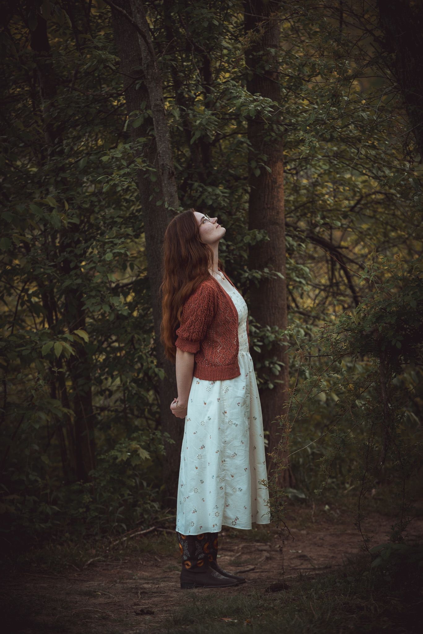 Figure in vintage dress and cardigan looking upward while standing on a forest path.