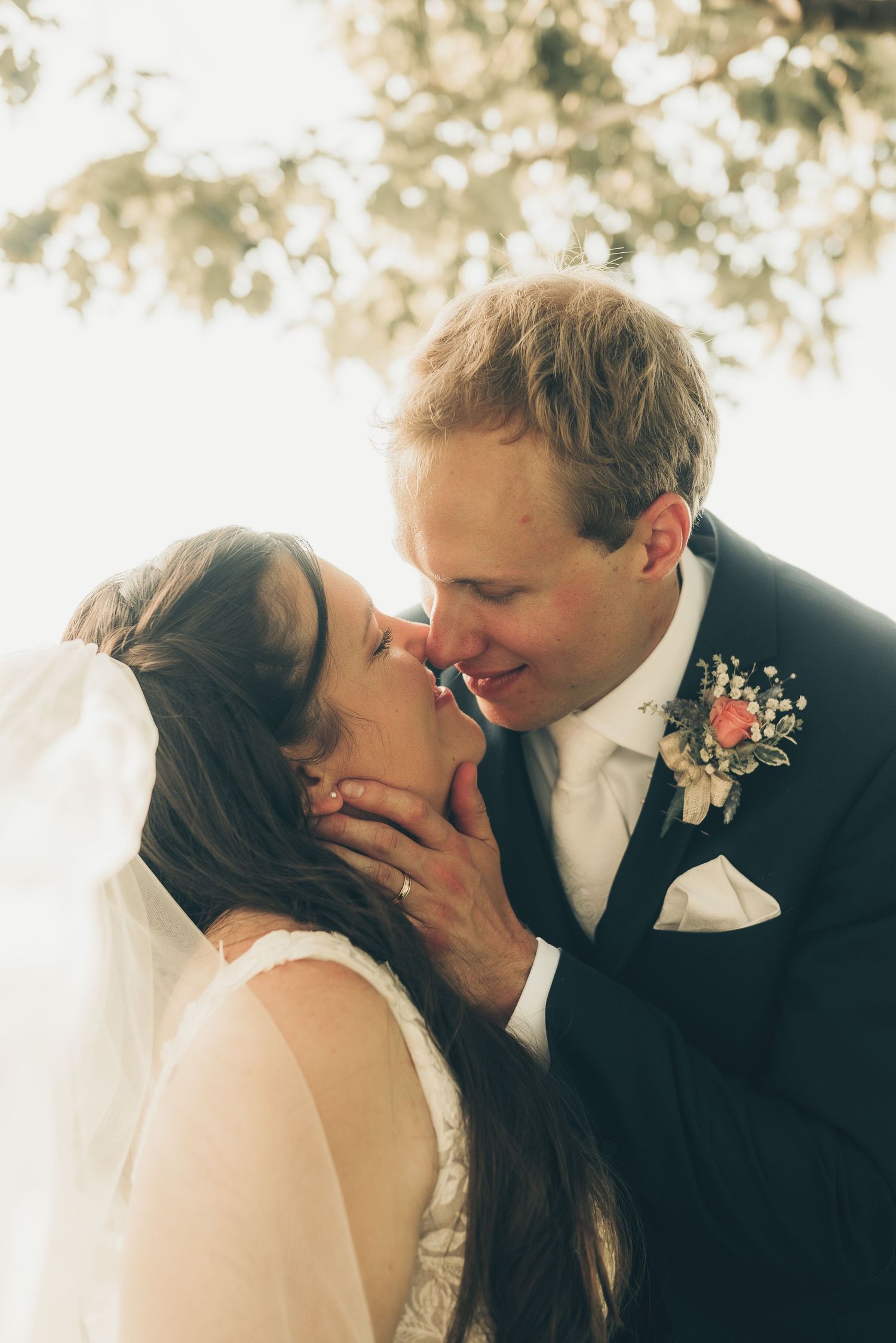 Bride and groom share a romantic almost-kiss, groom in dark suit with floral boutonniere, bride in white veil outdoors.
