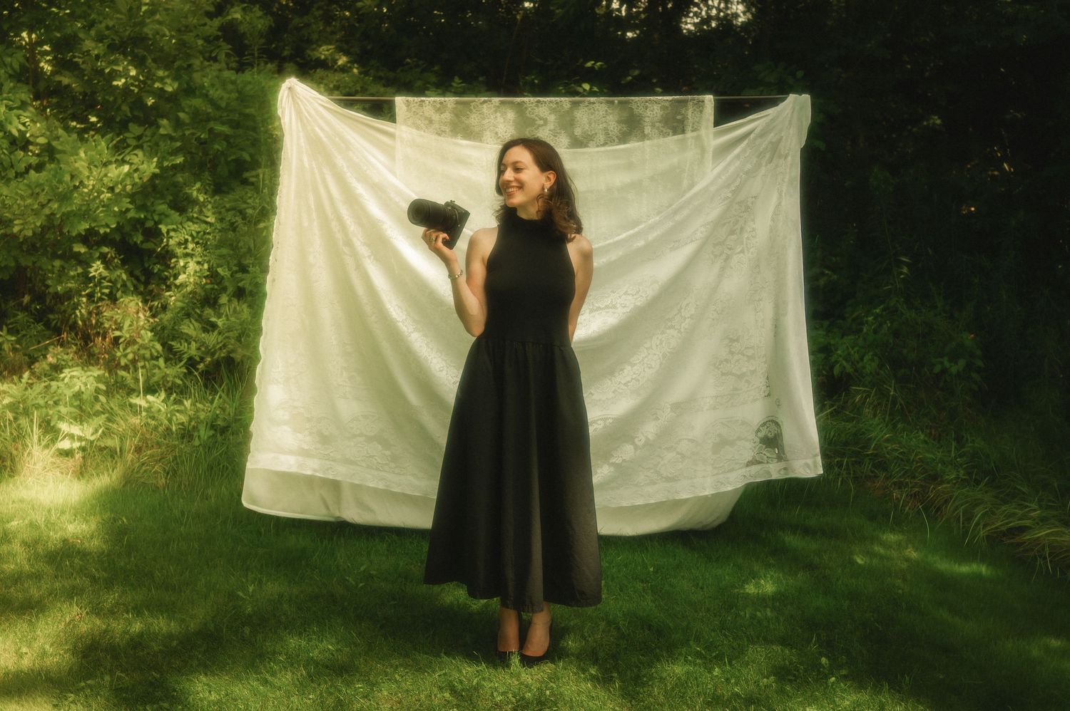 Woman in black dress holding camera stands smiling on grass before white sheet backdrop outdoors.