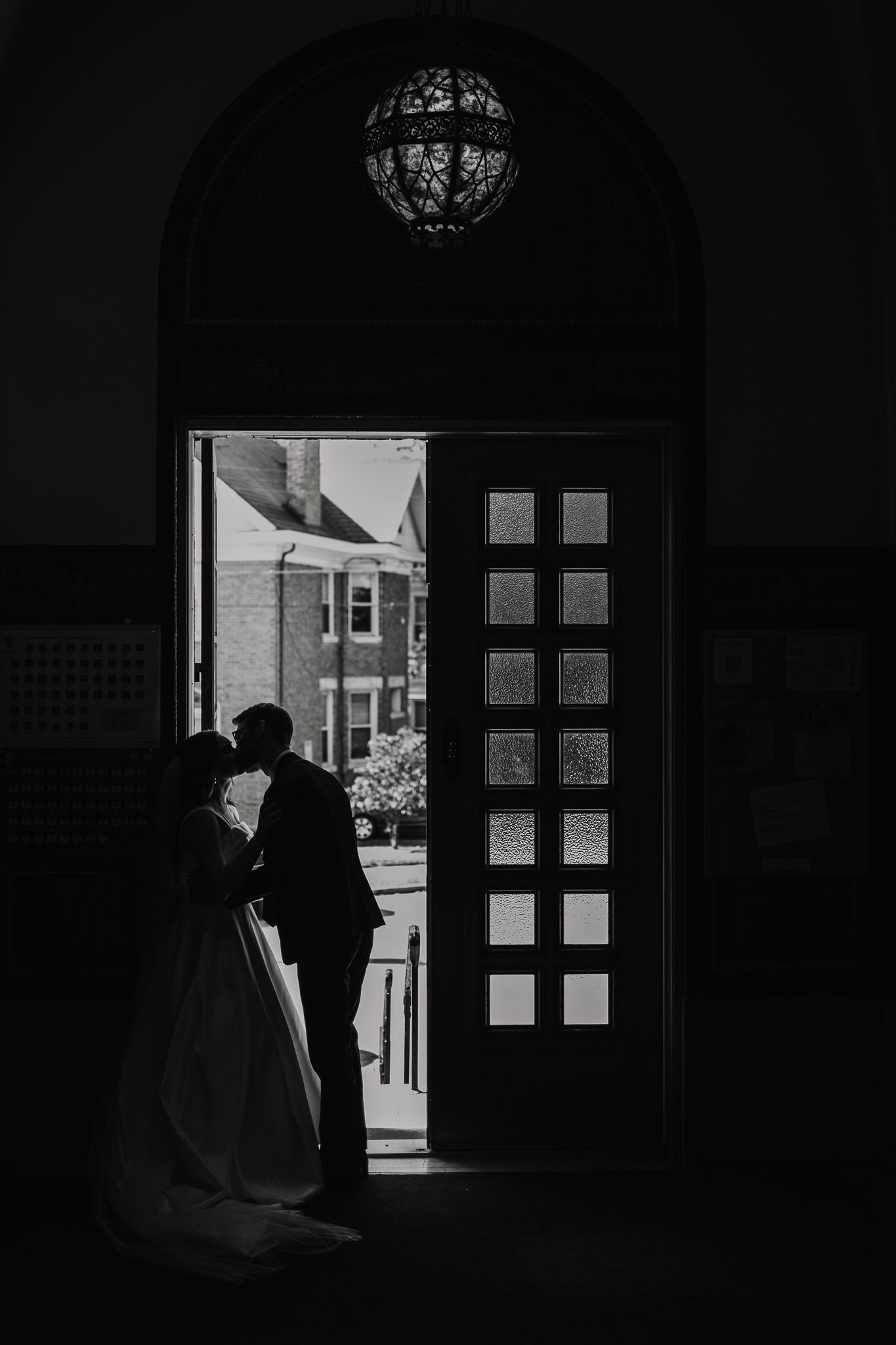 Silhouette of bride and groom kissing in arched doorway with decorative pendant light, black and white wedding photo.