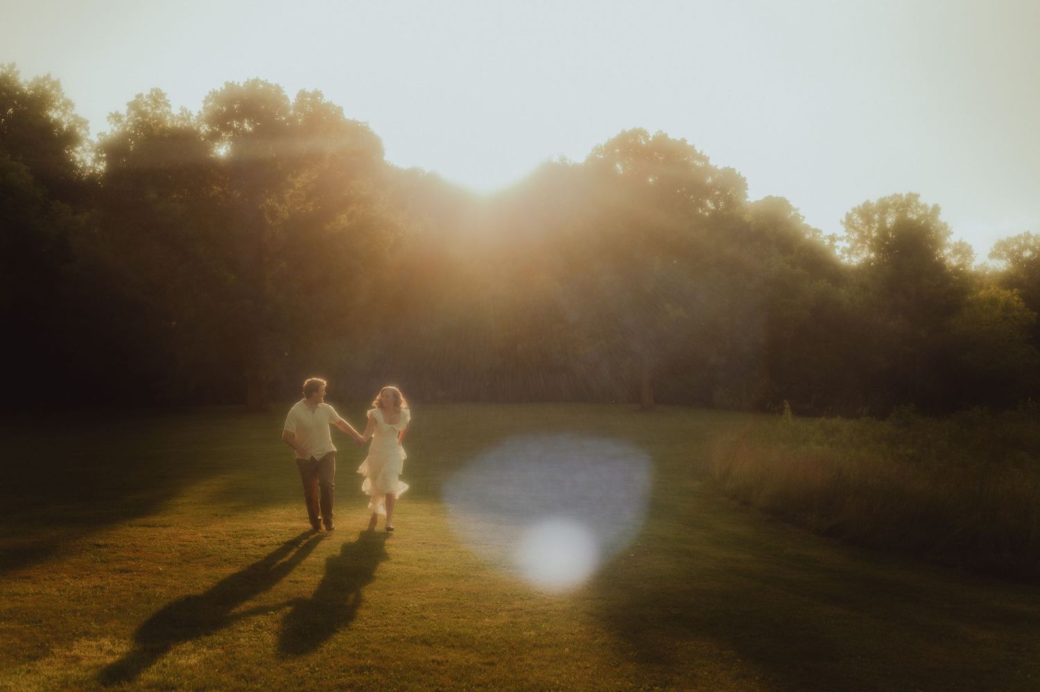 Two children run hand-in-hand across a sunlit green meadow surrounded by trees at golden hour.