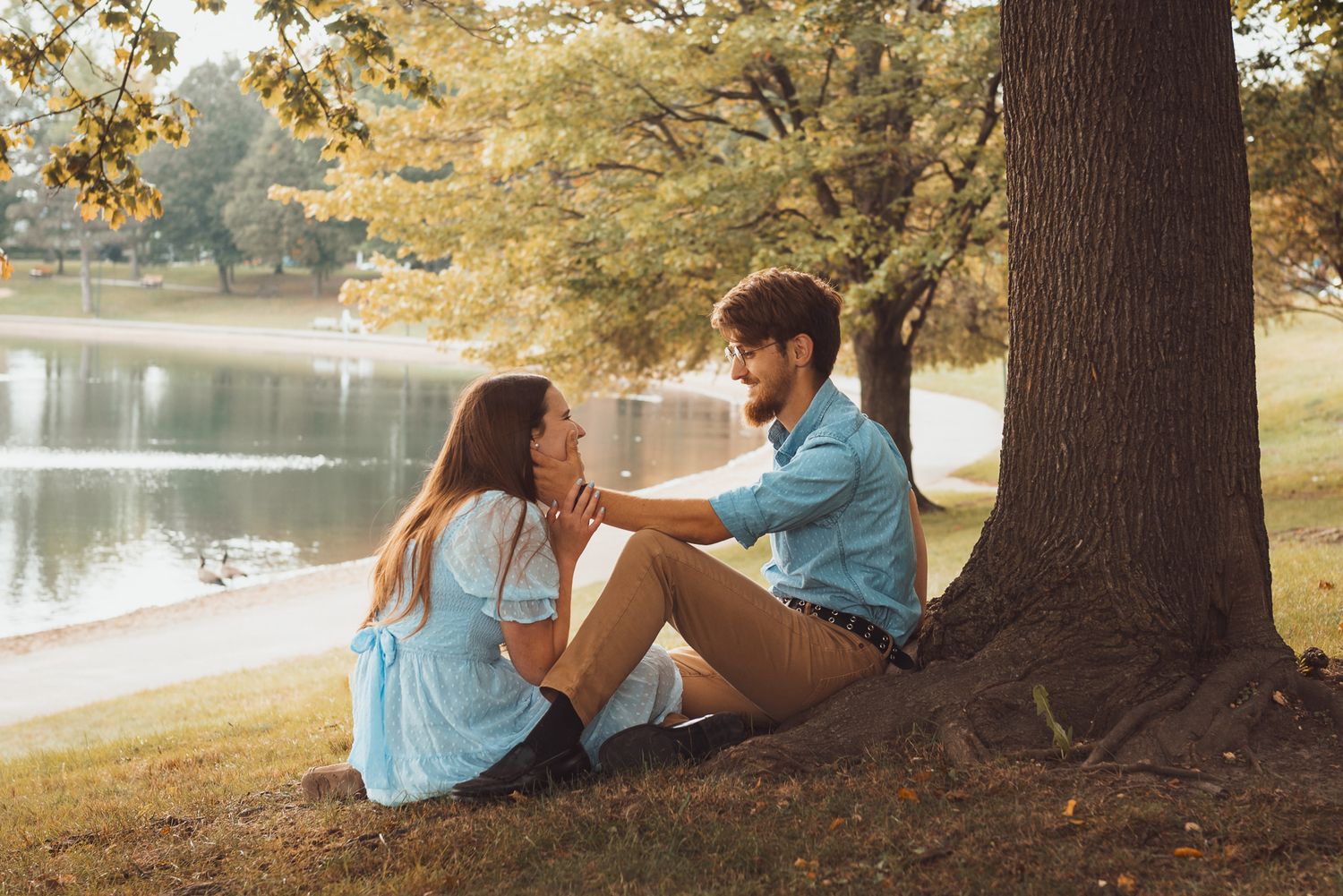A couple relaxes together under large trees by a peaceful lakeside setting.