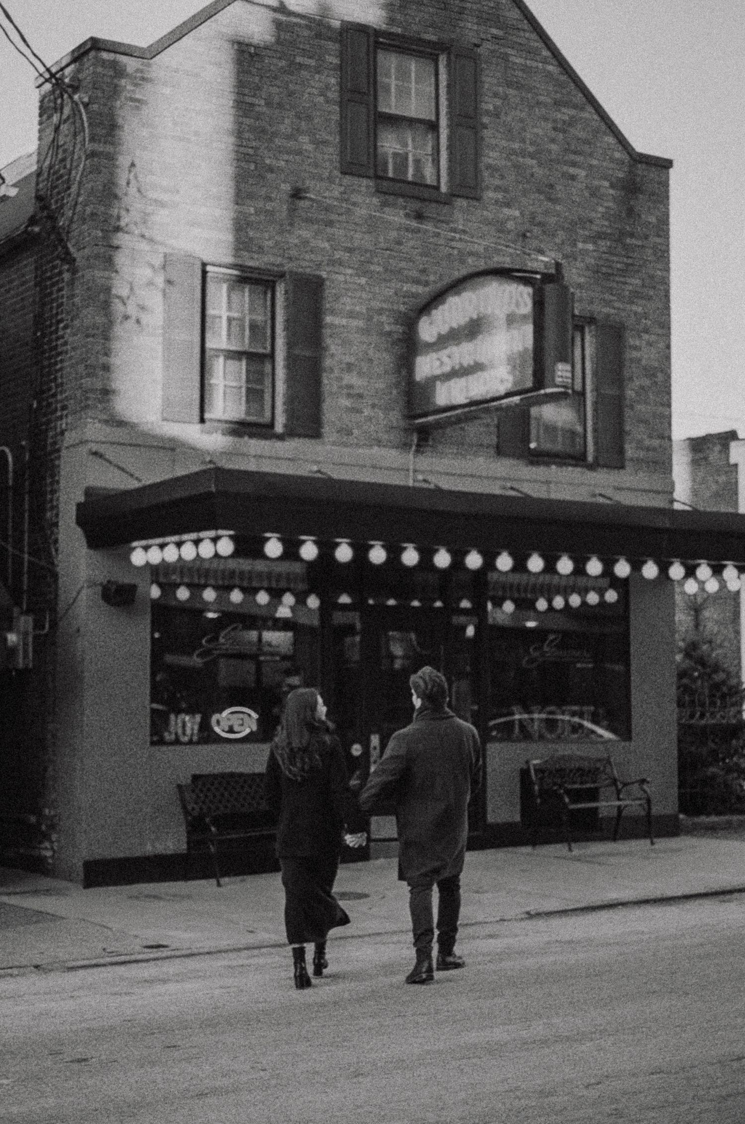 A black and white photograph of a vintage storefront with string lights and people walking on the sidewalk in the 1950s.