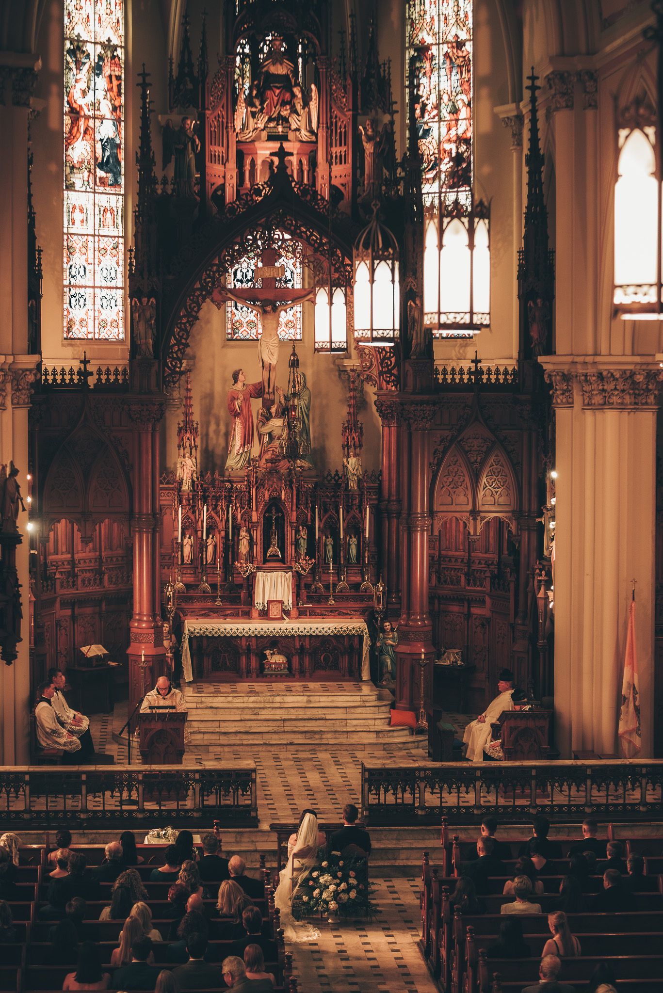 Interior of a Gothic cathedral during a wedding ceremony with stained glass windows and wooden pews.