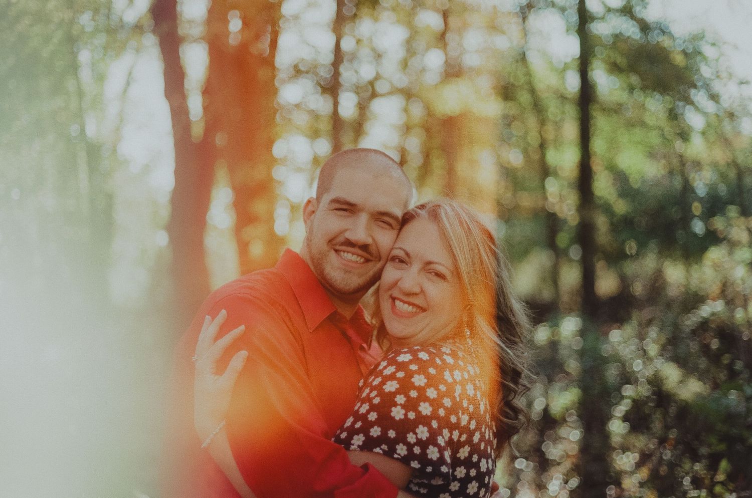 Couple embraces in a sunlit forest with warm orange glow filtering through trees during golden hour.
