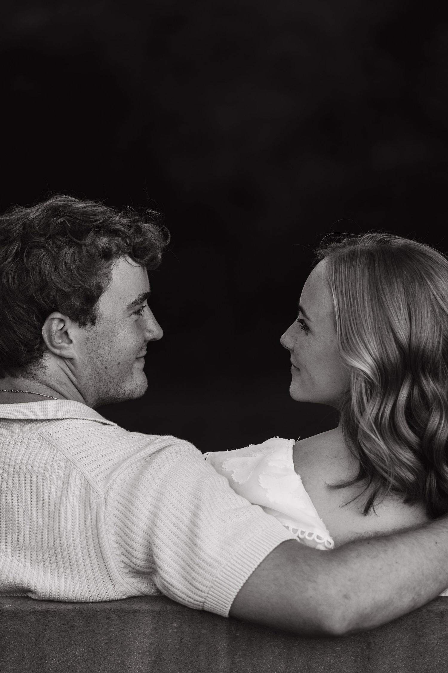 Black and white photo of a romantic couple gazing at each other, woman's arm around man's shoulder.