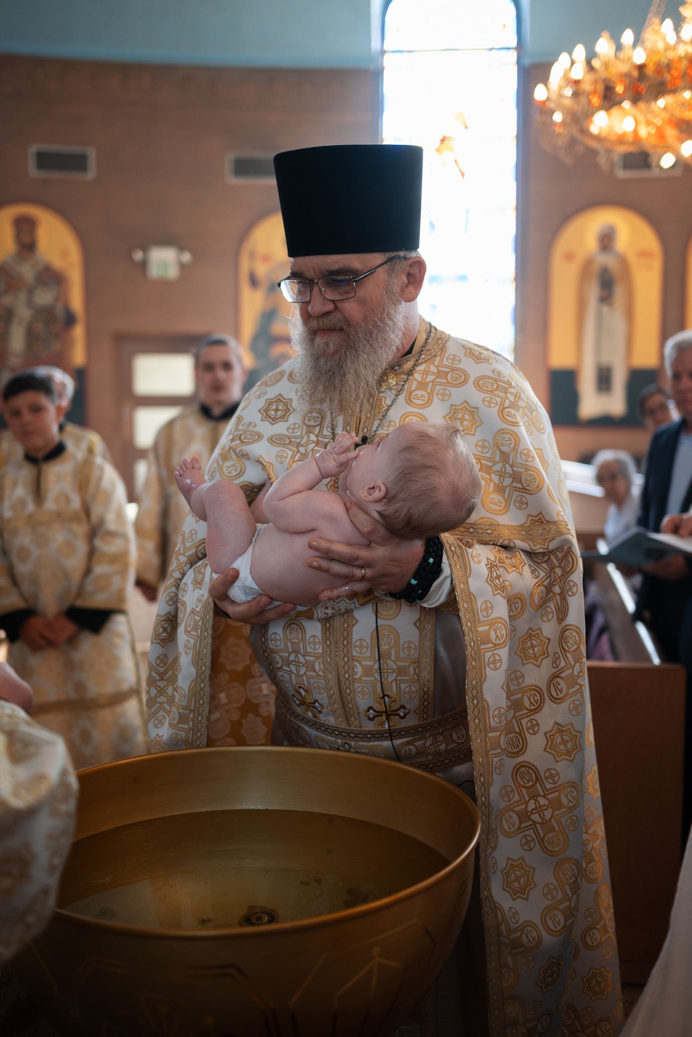 Orthodox priest in gold vestments performs baptism ceremony with infant in ornate church interior.