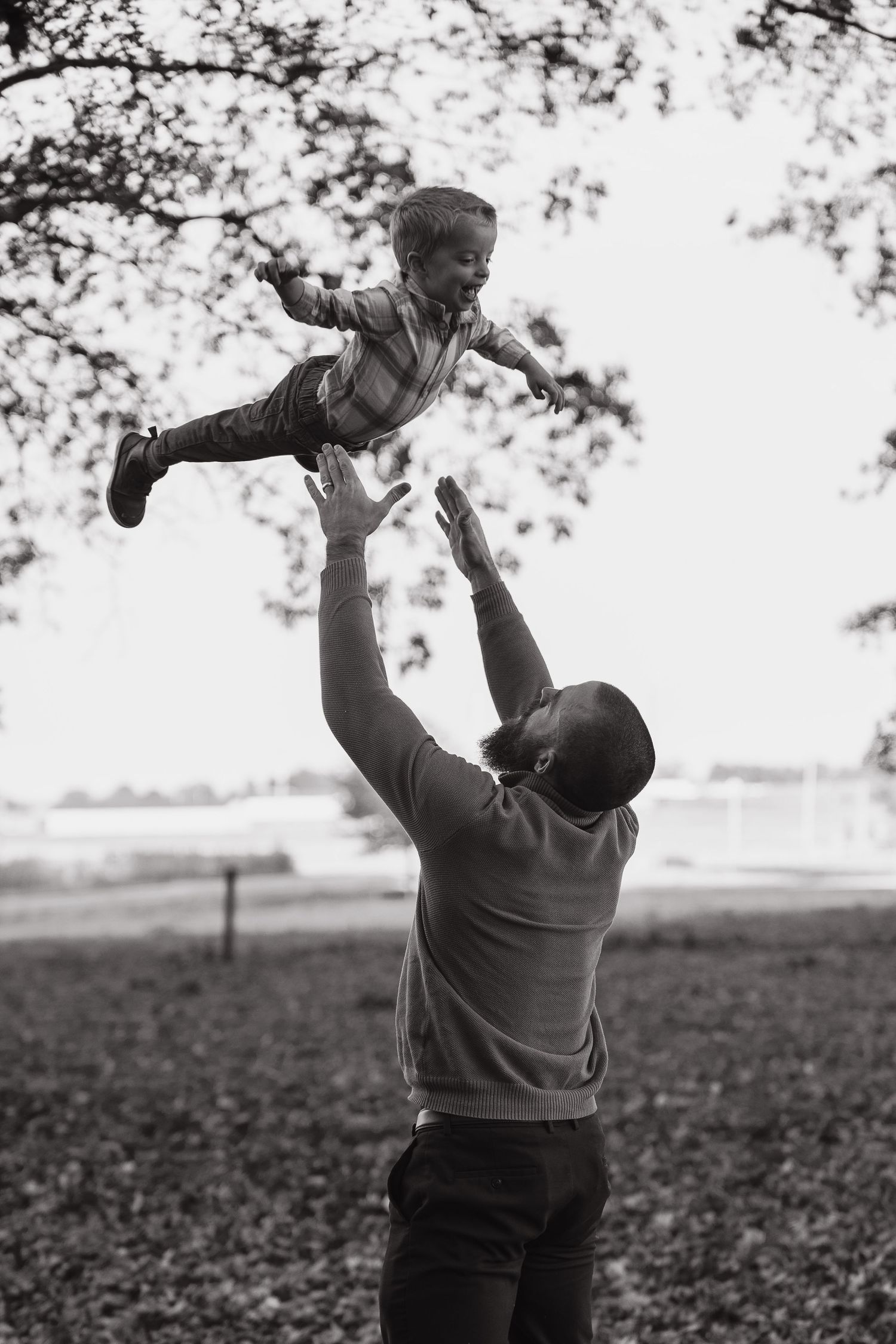 Black and white photo of a father joyfully tossing his young child into the air in an outdoor park setting.