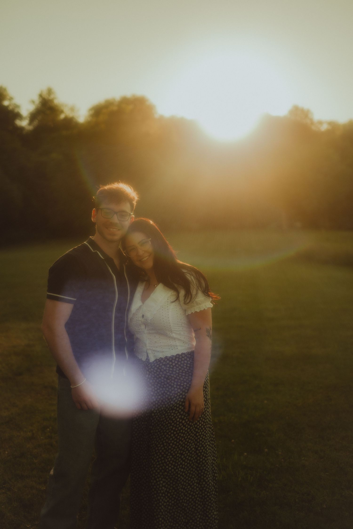 Couple embracing at golden hour sunset in a lush green park, warm lens flare glowing behind them.