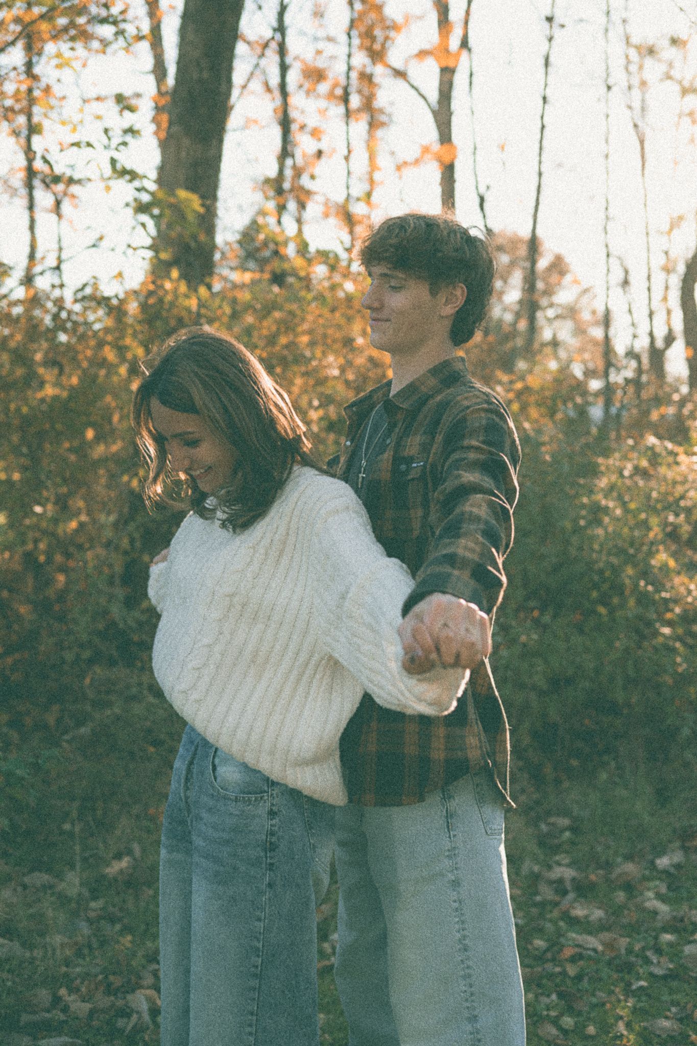 Two people in jeans and sweaters embrace while standing among autumn trees with orange foliage.