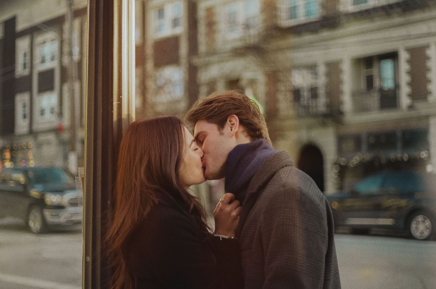 A couple shares a romantic kiss on a city street with urban buildings in the background.
