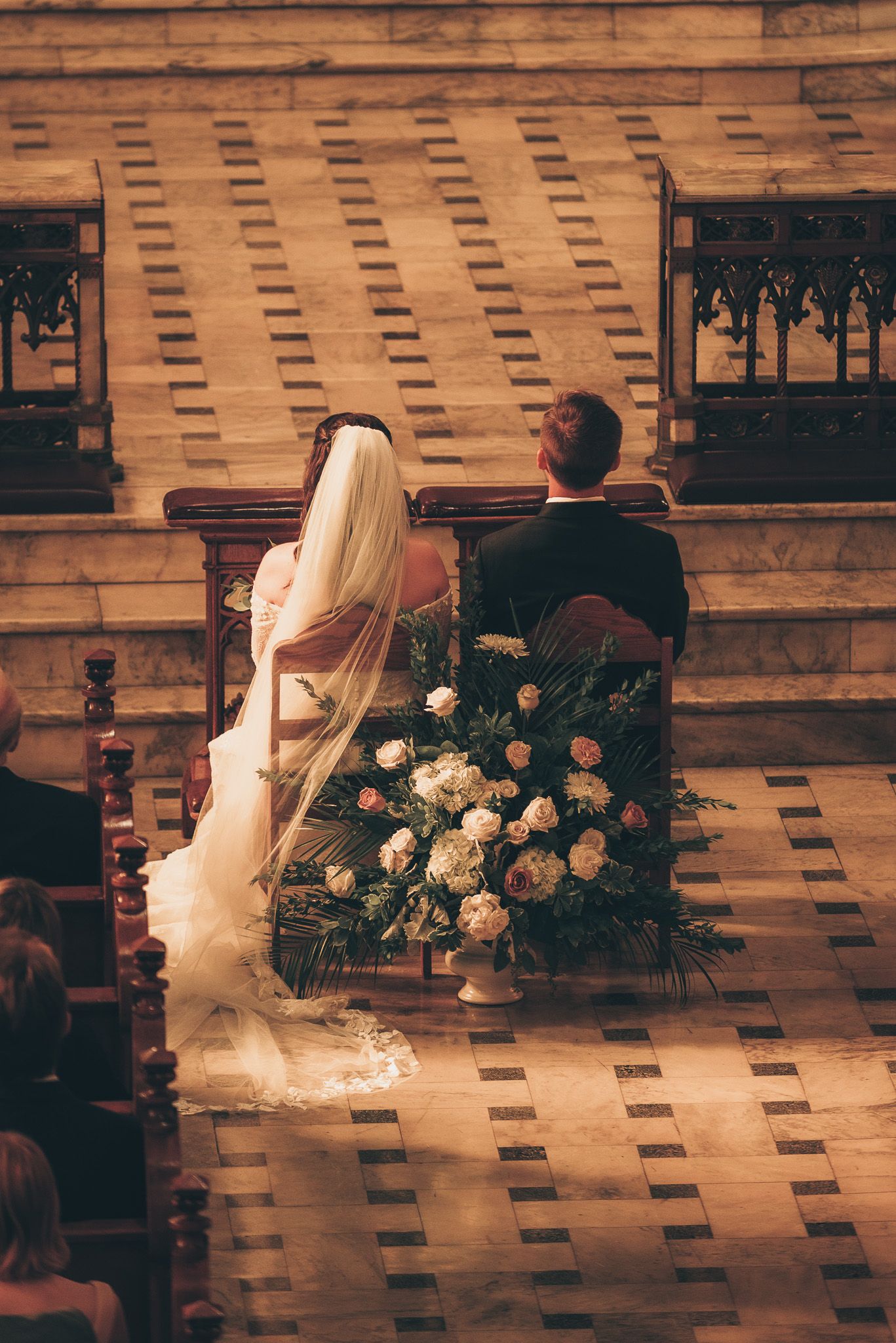 Wedding couple seated in church pews with floral arrangements during ceremony.