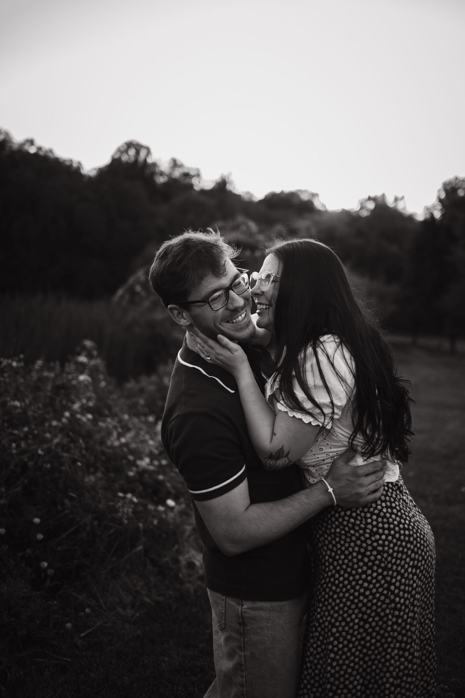 Black and white photo of a couple embracing and laughing together outdoors at dusk near lush greenery.