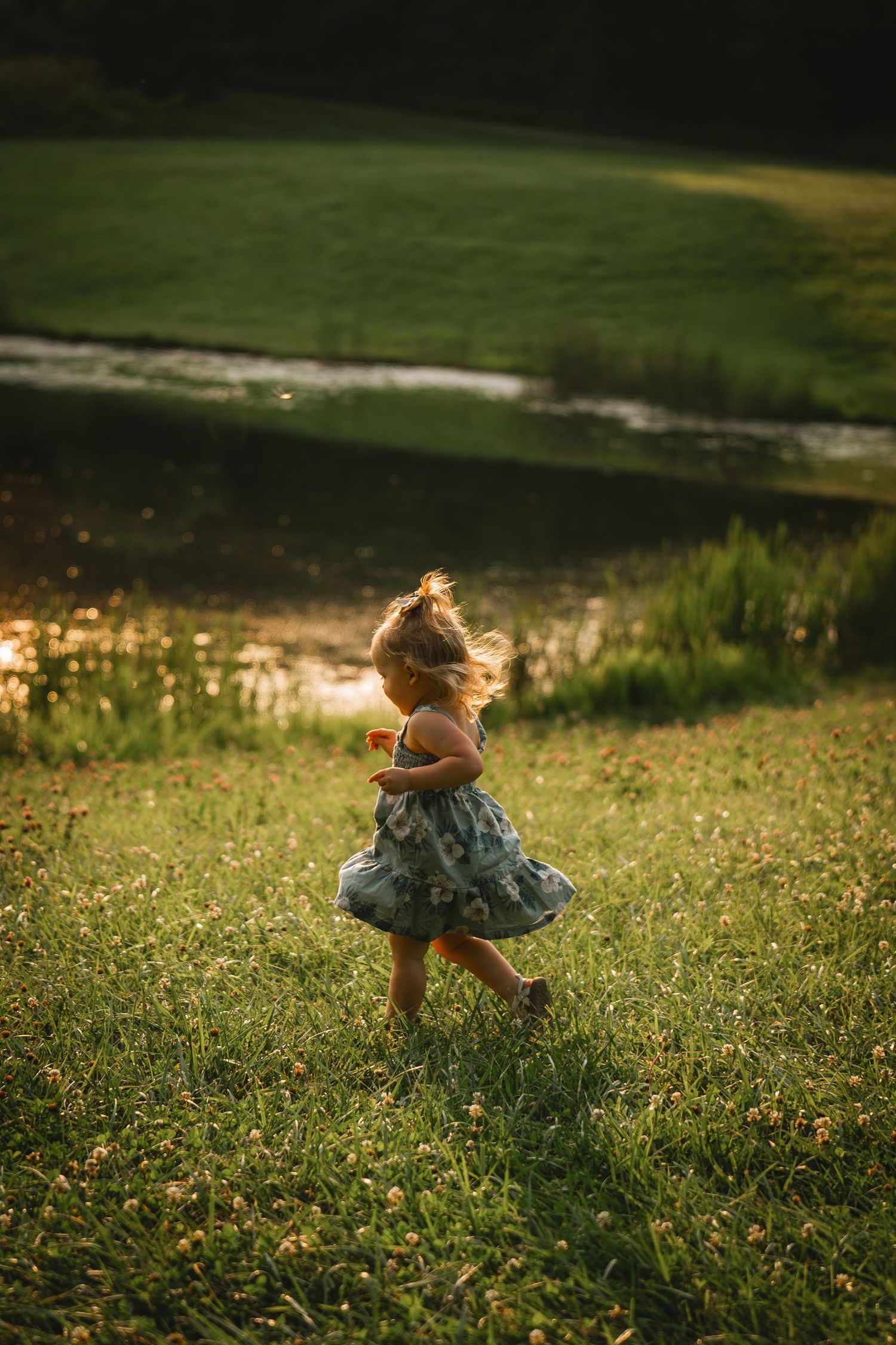 Young girl in floral dress running through sunlit green meadow near a peaceful stream at golden hour.