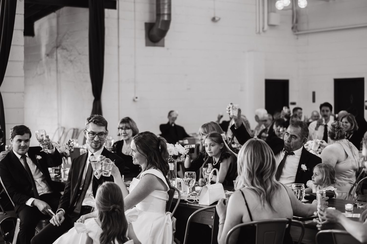Black and white wedding reception toast with guests raising glasses in an elegant venue.