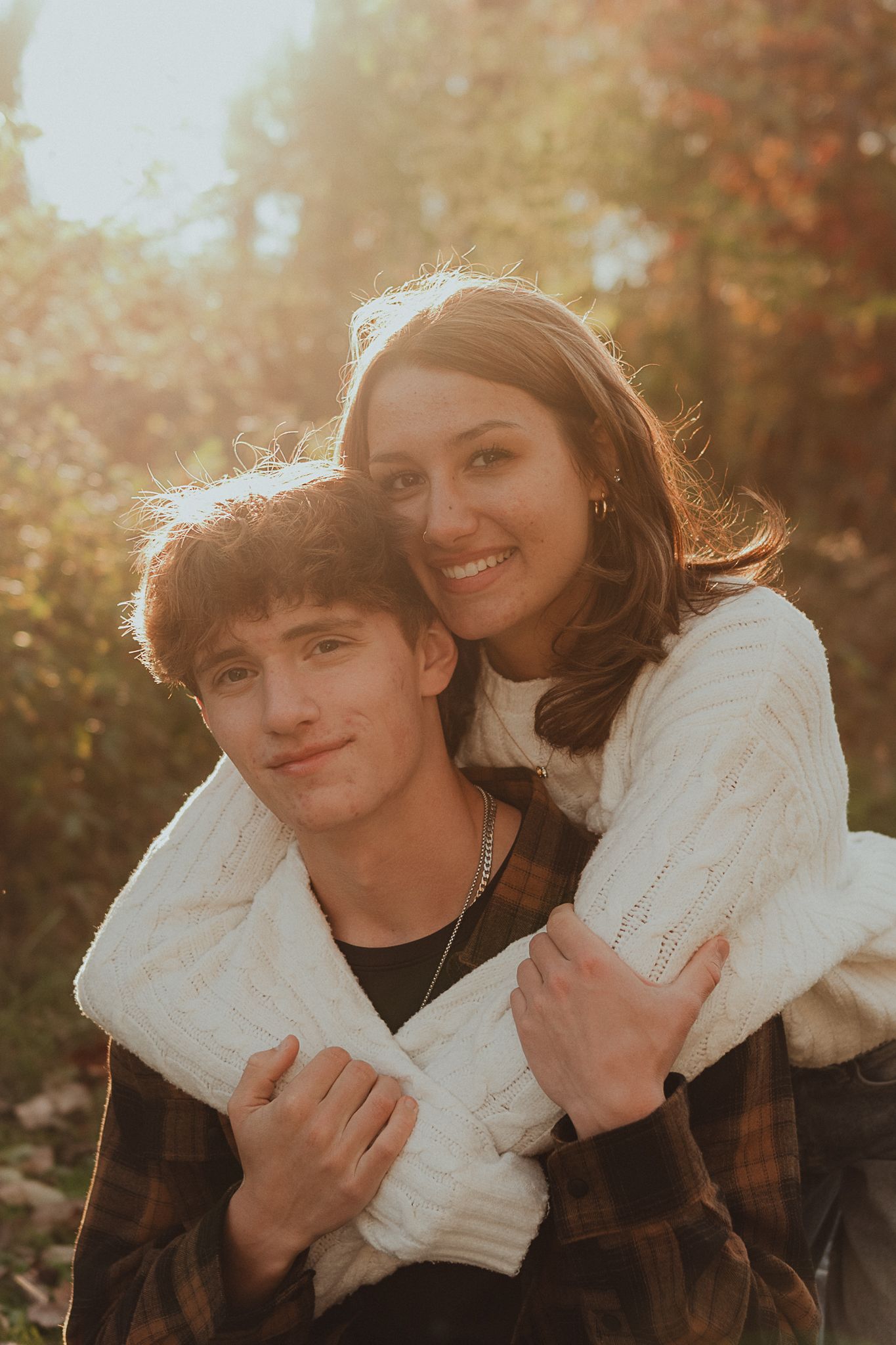 A couple embraces outdoors during golden hour sunlight with autumn foliage in the background.