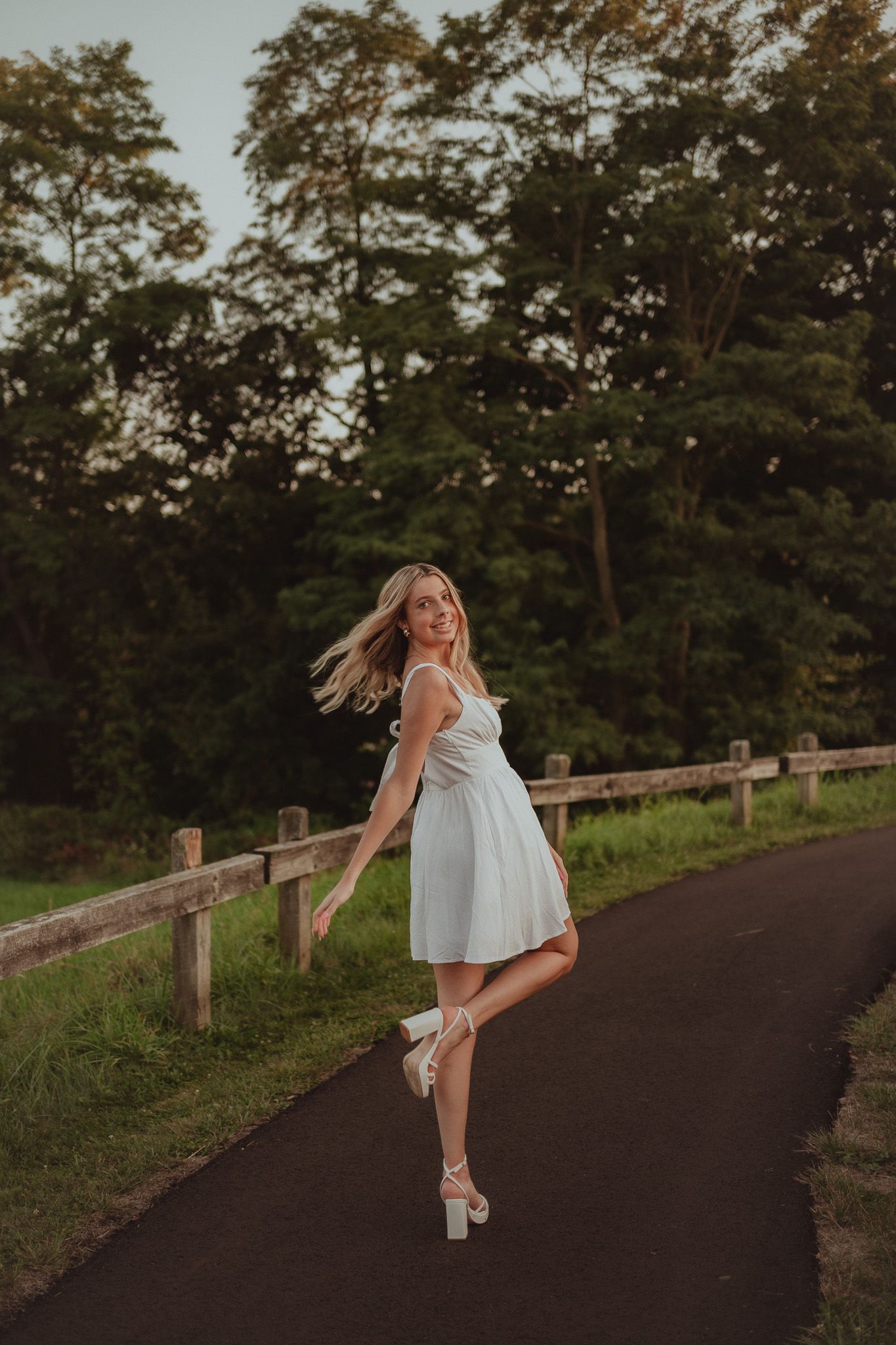 Person in a white dress twirling playfully on a path with a wooden fence and trees in the background.