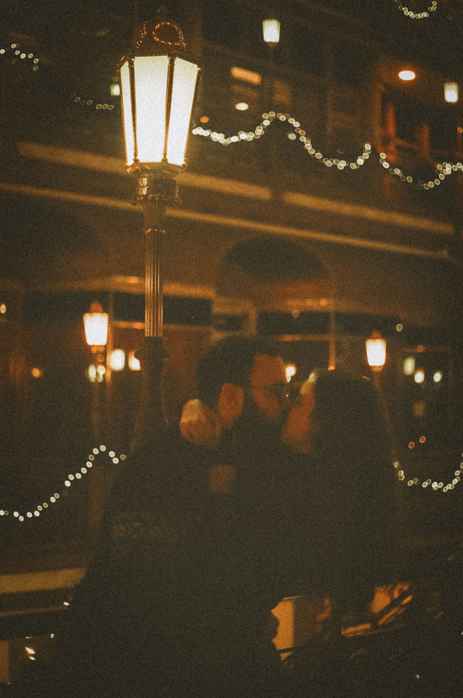 Vintage-style street lamp glows warmly against a dark evening scene with decorative string lights and blurred figures.