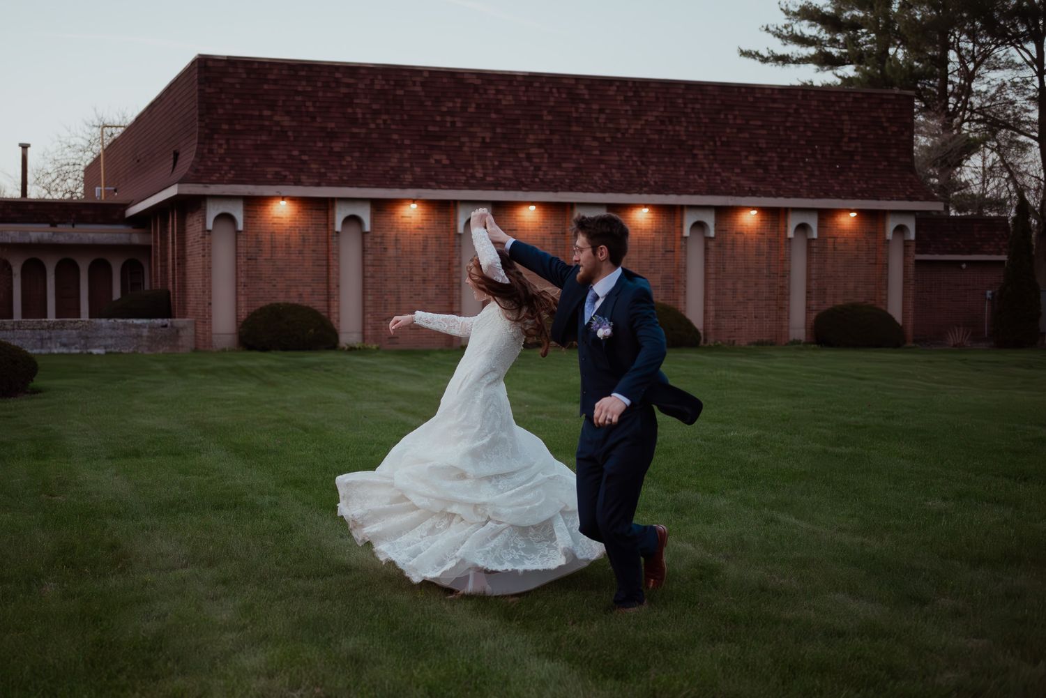 A wedding couple poses for romantic photos on a grassy lawn near a brick building at dusk.