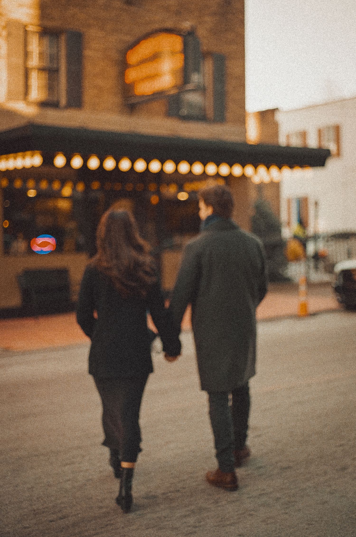 Two people walking together on a snowy sidewalk at dusk with warm glowing lights in the background.