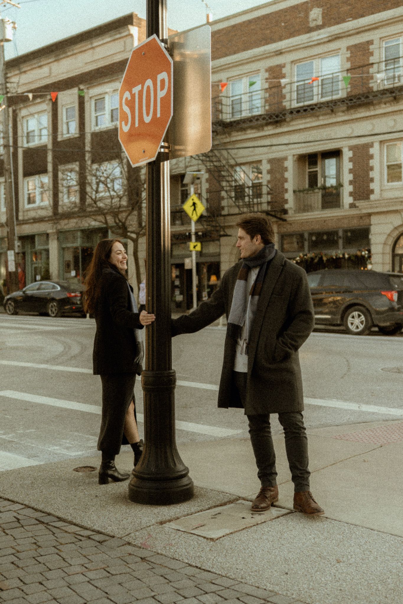Two people stand chatting by a vintage-style lamppost with a stop sign on a city street corner.