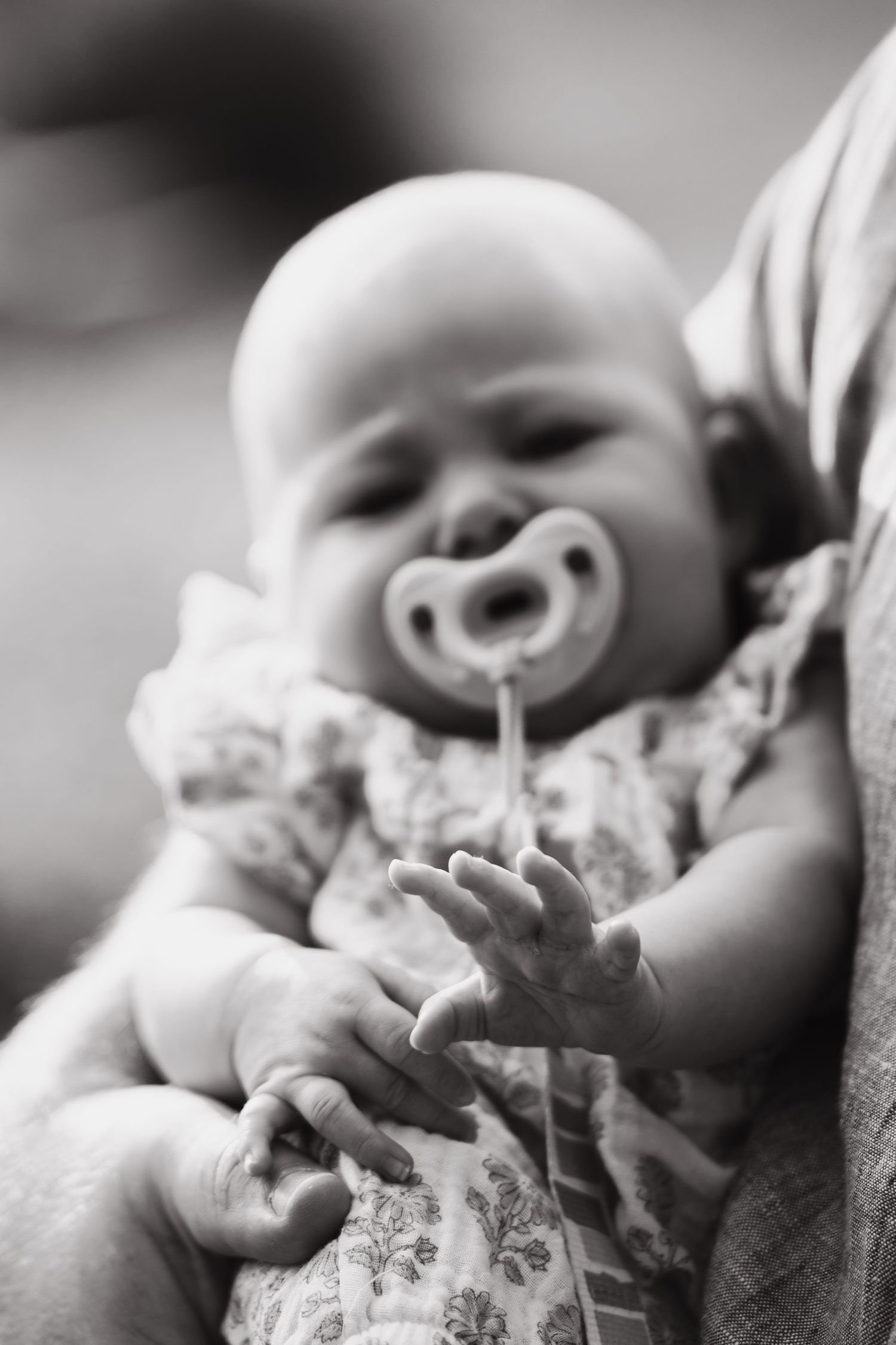 Black and white photo of a peaceful baby with a pacifier, cradled in an adult's arms, wearing a floral outfit.