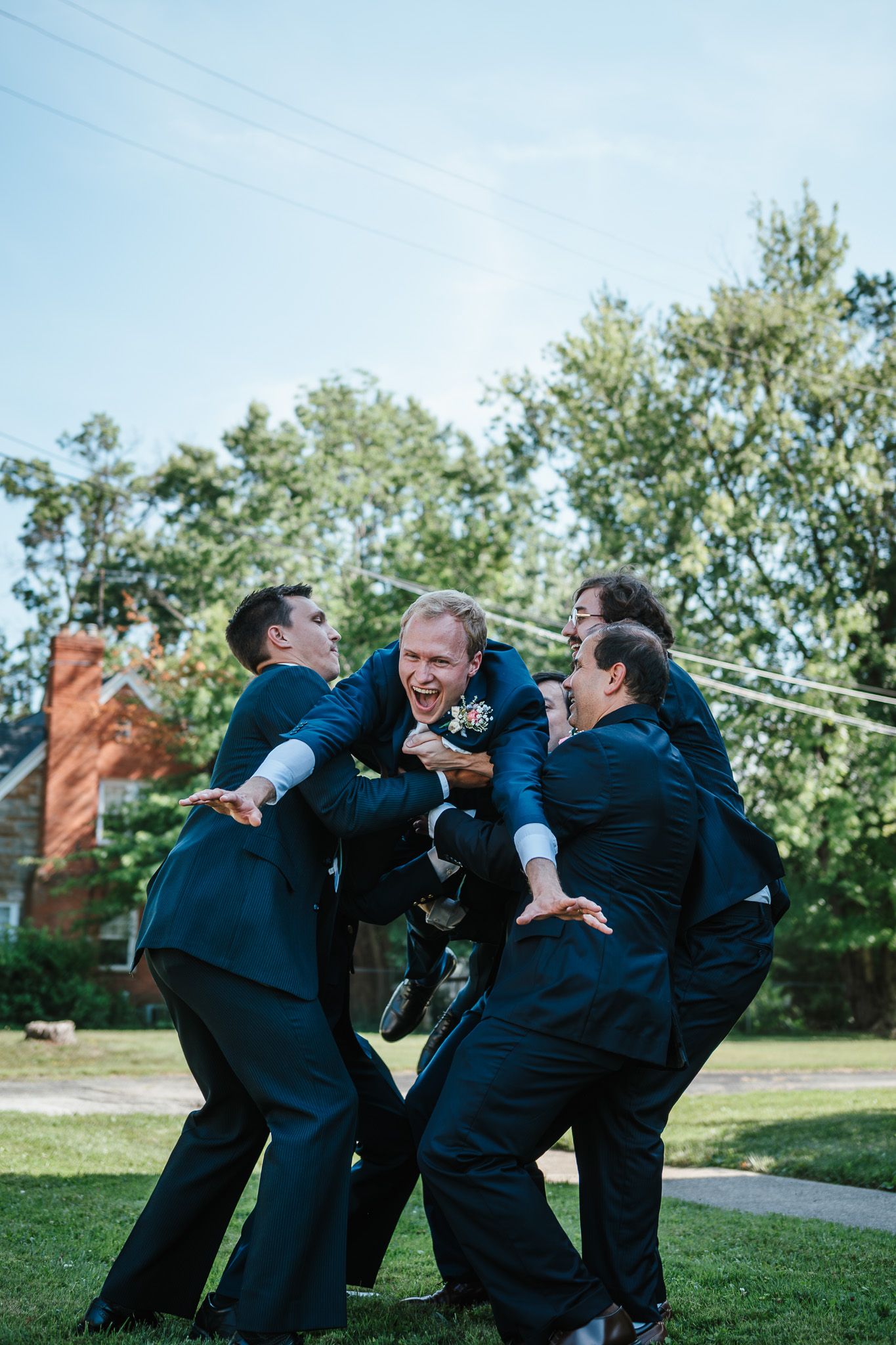 Groomsmen in dark suits playfully pose and interact on grassy lawn during wedding photo session.