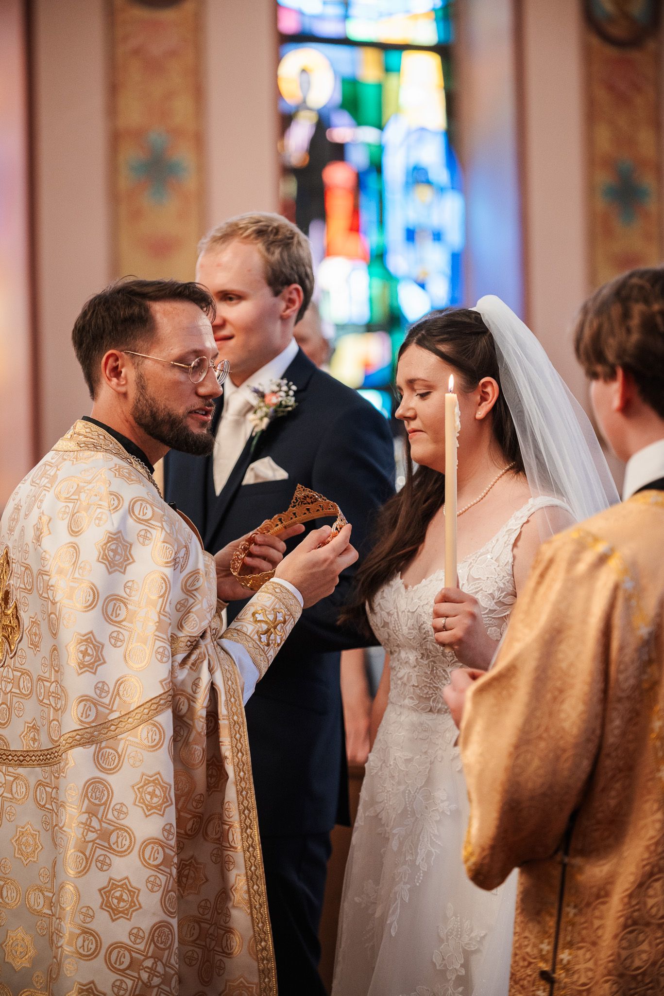Orthodox priests in ornate gold vestments perform traditional wedding ceremony rituals in church with stained glass windows.