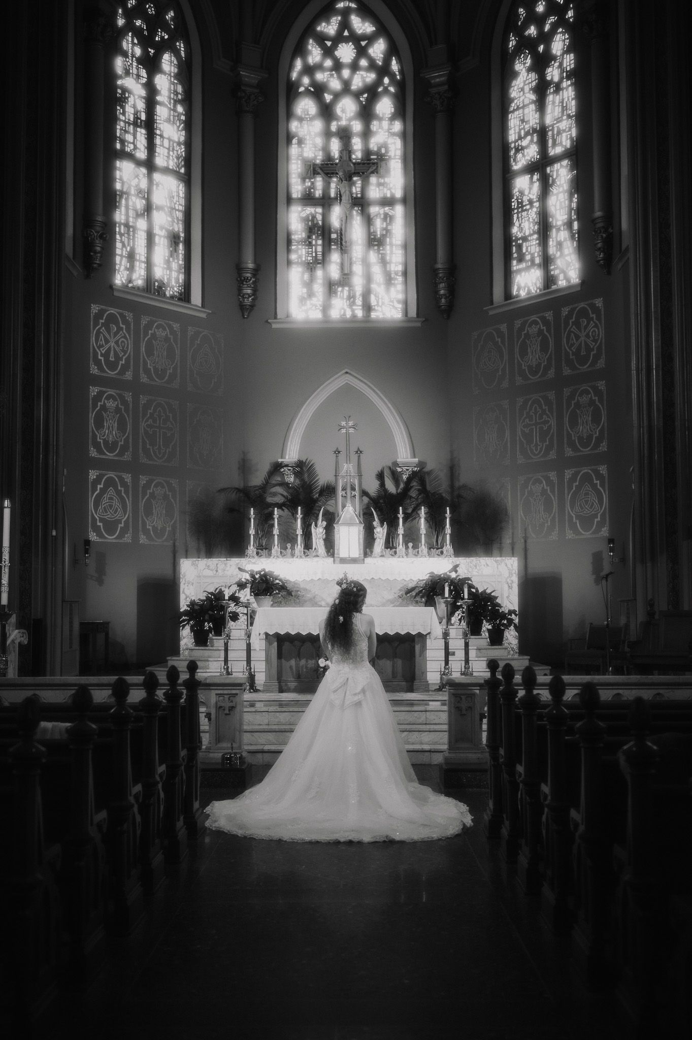 Black and white photo of a bride in a white gown standing at a church altar with stunning stained glass windows.