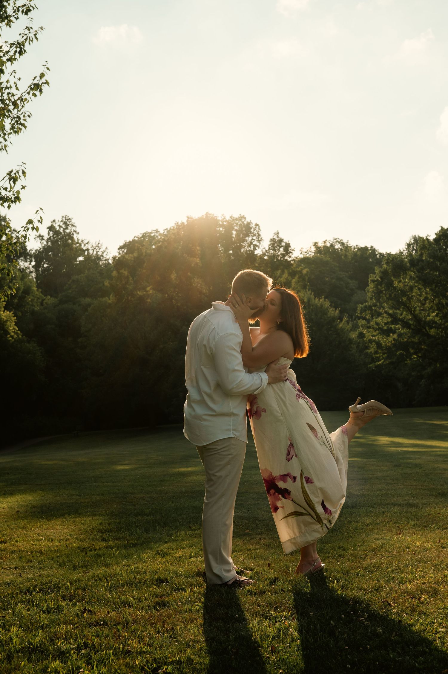 Couple sharing a romantic kiss at golden hour in a lush green park, woman wearing a floral dress.