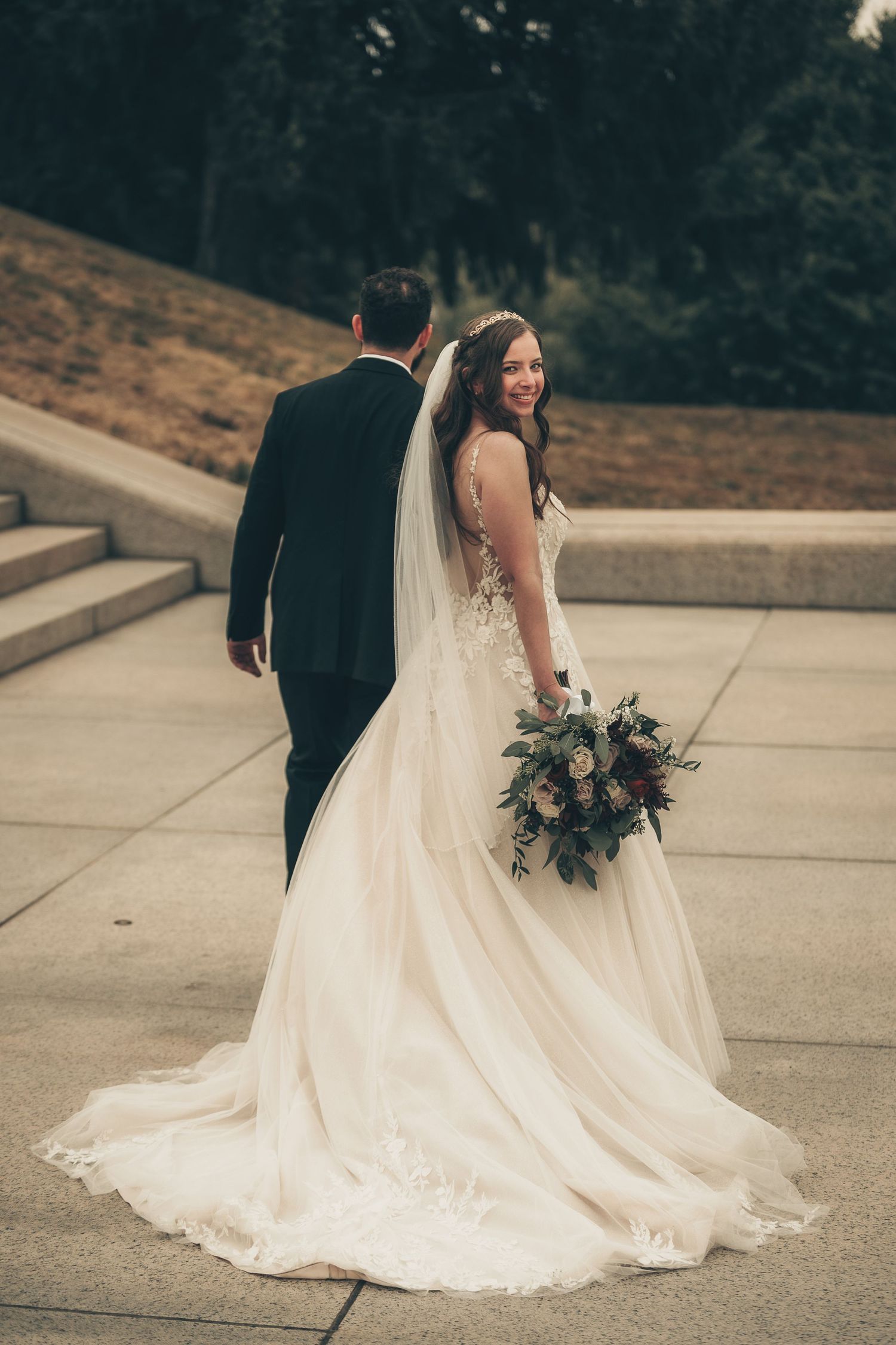 Bride in white gown with veil holding bouquet walks with groom in black suit on stone pathway outdoors.