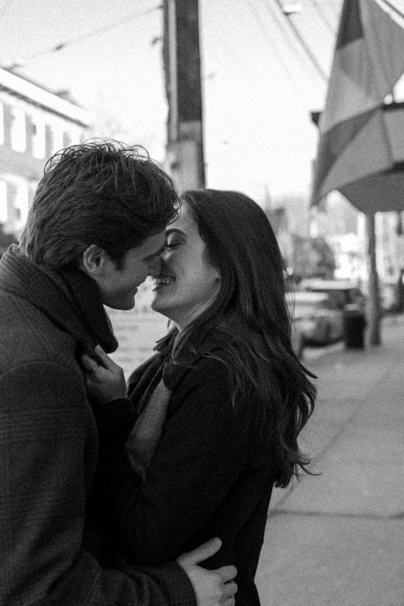 A romantic black and white street scene of a couple in dark coats sharing an intimate moment on the sidewalk.
