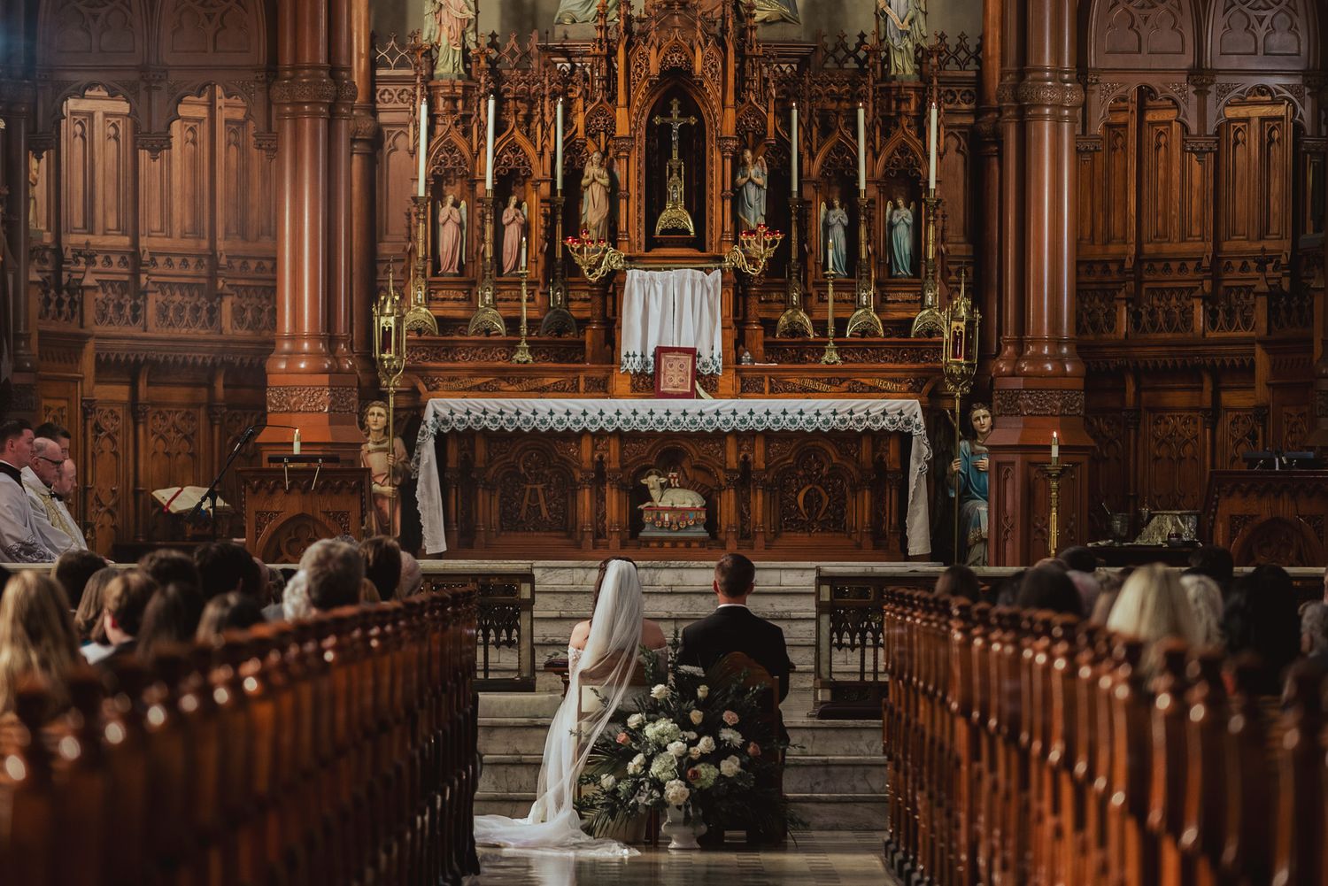 A couple kneels at the altar of an ornate Catholic church during a traditional wedding ceremony.