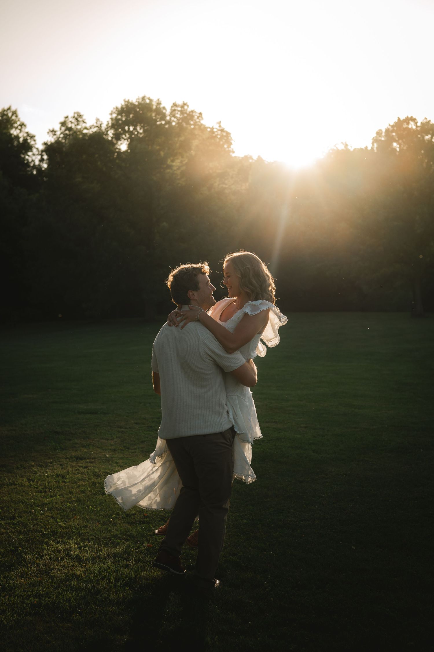 Couple embracing at golden hour sunset in lush green field, man lifting woman in white dress.