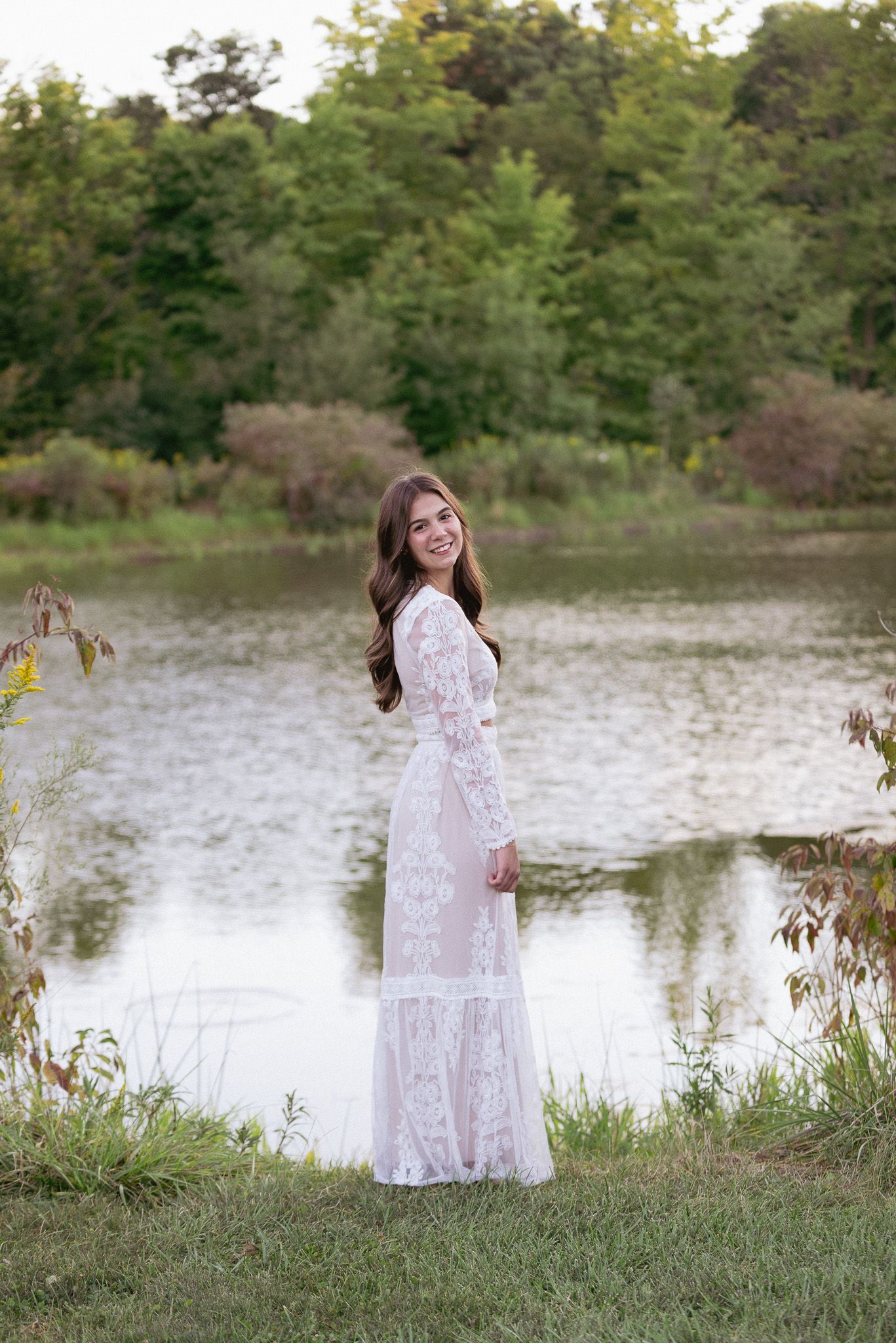 A woman in a lace gown poses by a serene lake during golden hour.