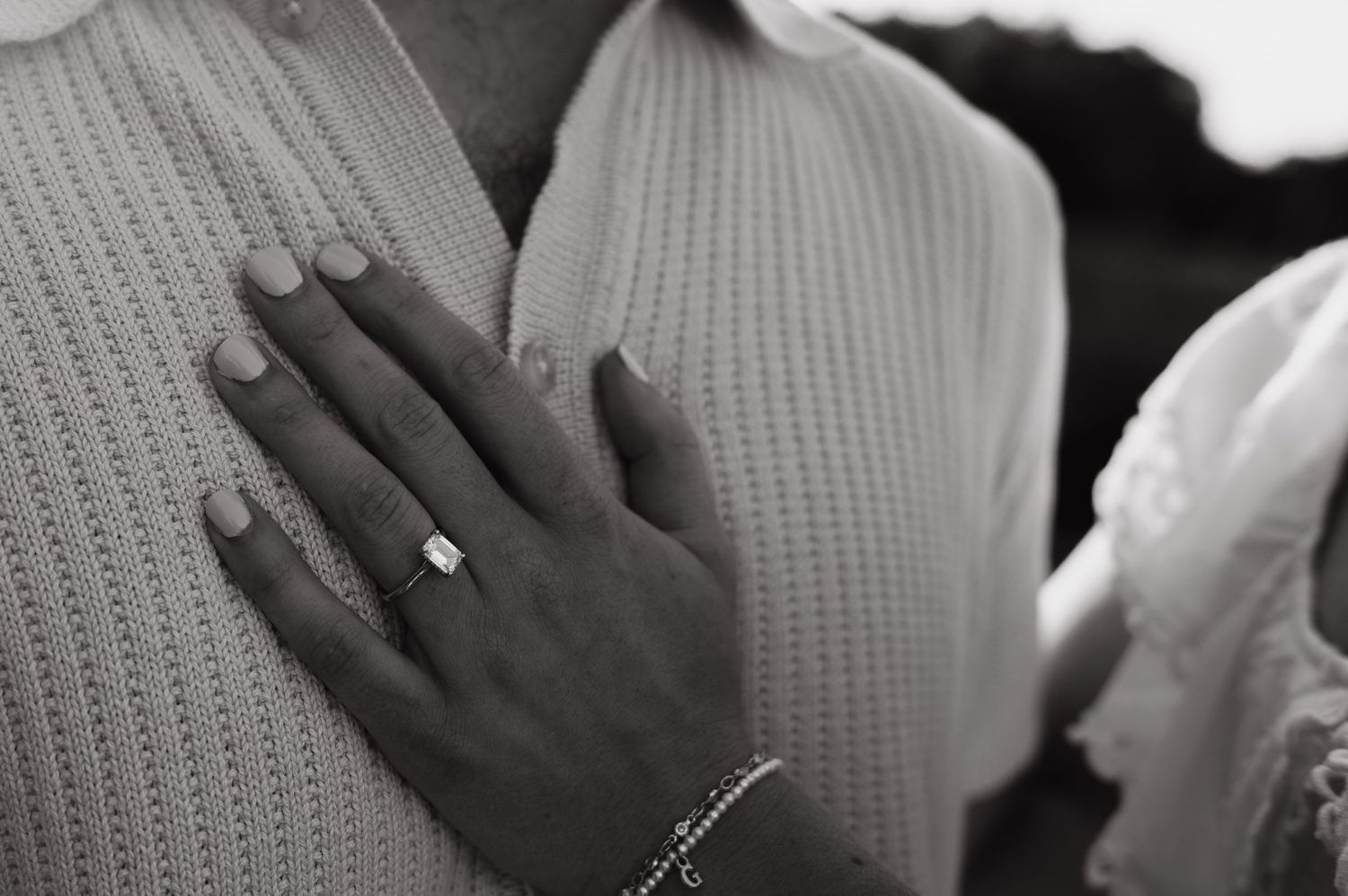 Black and white photo of a woman's hand with an engagement ring resting on a man's chest.