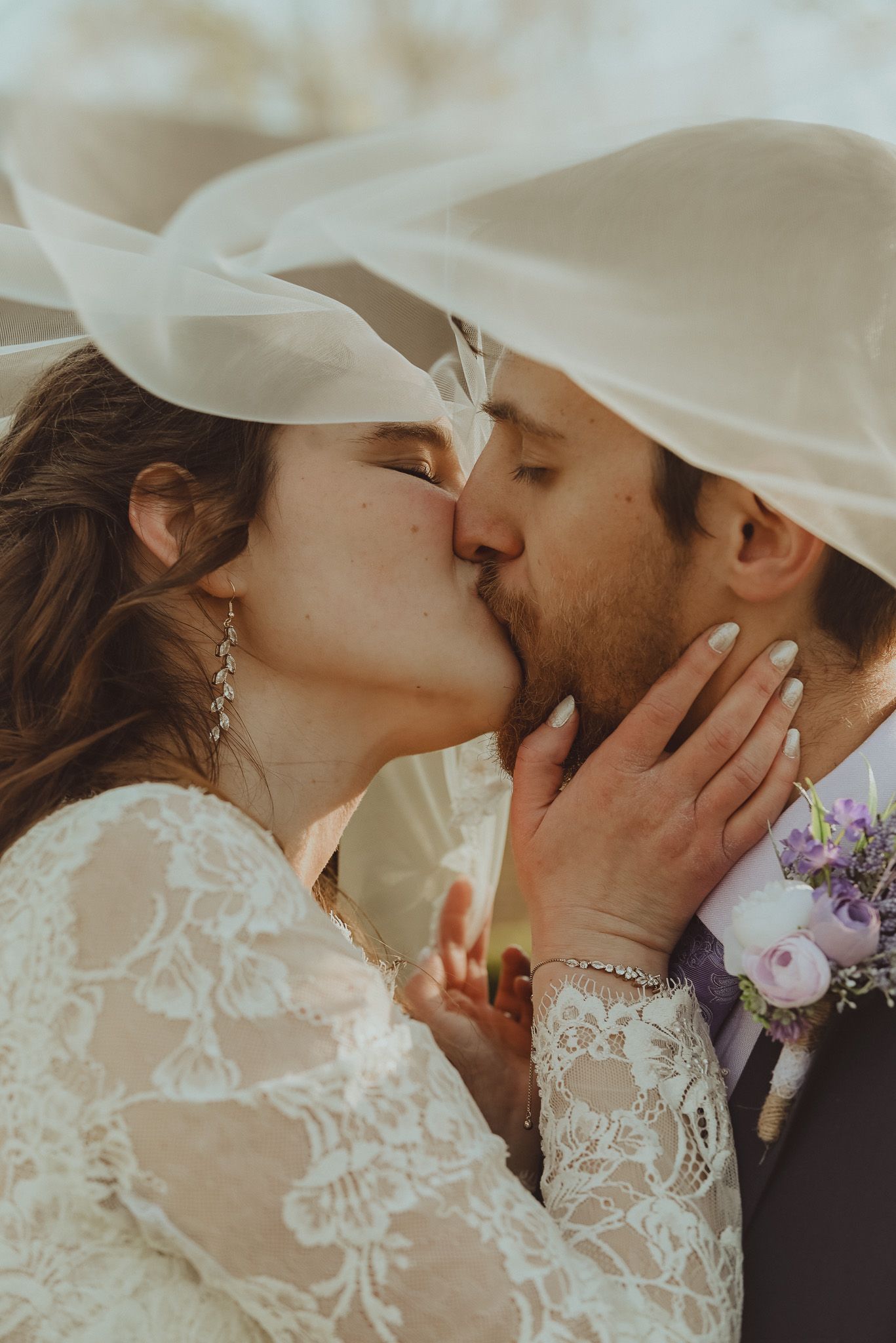 Intimate moment shared under a wedding veil with lace dress detail and floral boutonniere.